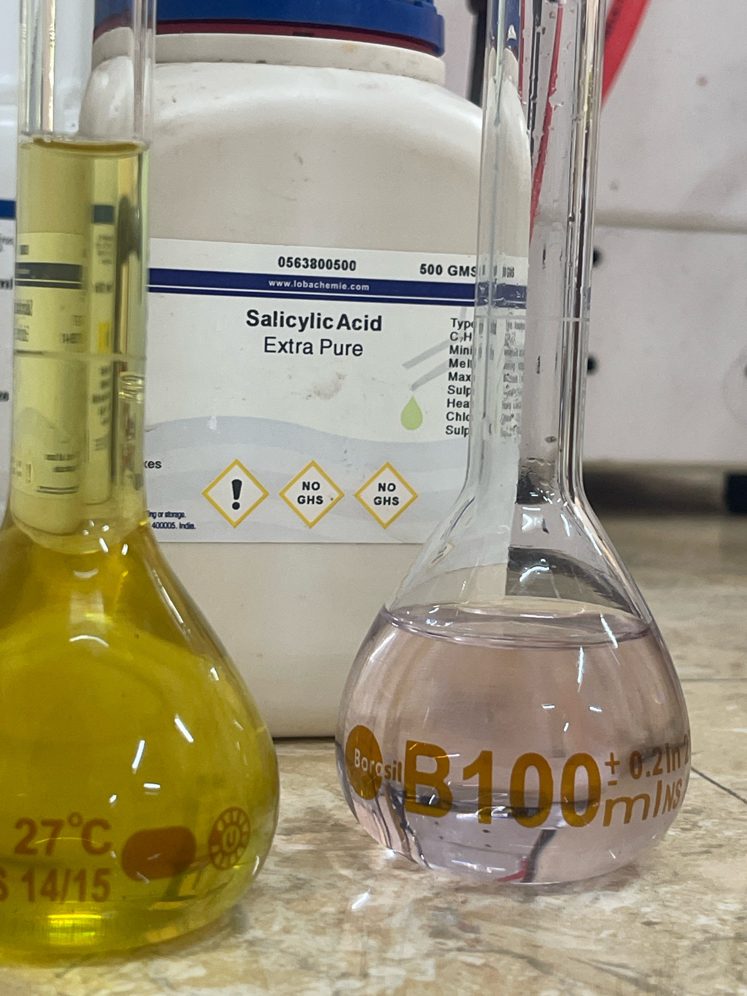 The image displays a laboratory setting with two flasks containing liquids. A large white container labeled "Salicylic Acid Extra Pure" is visible in the background, along with its chemical composition and hazard symbols. One flask on the left holds a yellow liquid, and the flask on the right contains a pinkish liquid. The flask on the right is marked with "Borosil" and "B100±0.2mlNS." The scene is lit by overhead lighting, and the flasks rest on a tiled surface. The location is identified as Khankar Gaon, India, though there is no visual information in the image to confirm this geographical placement. No individuals are present in the image.