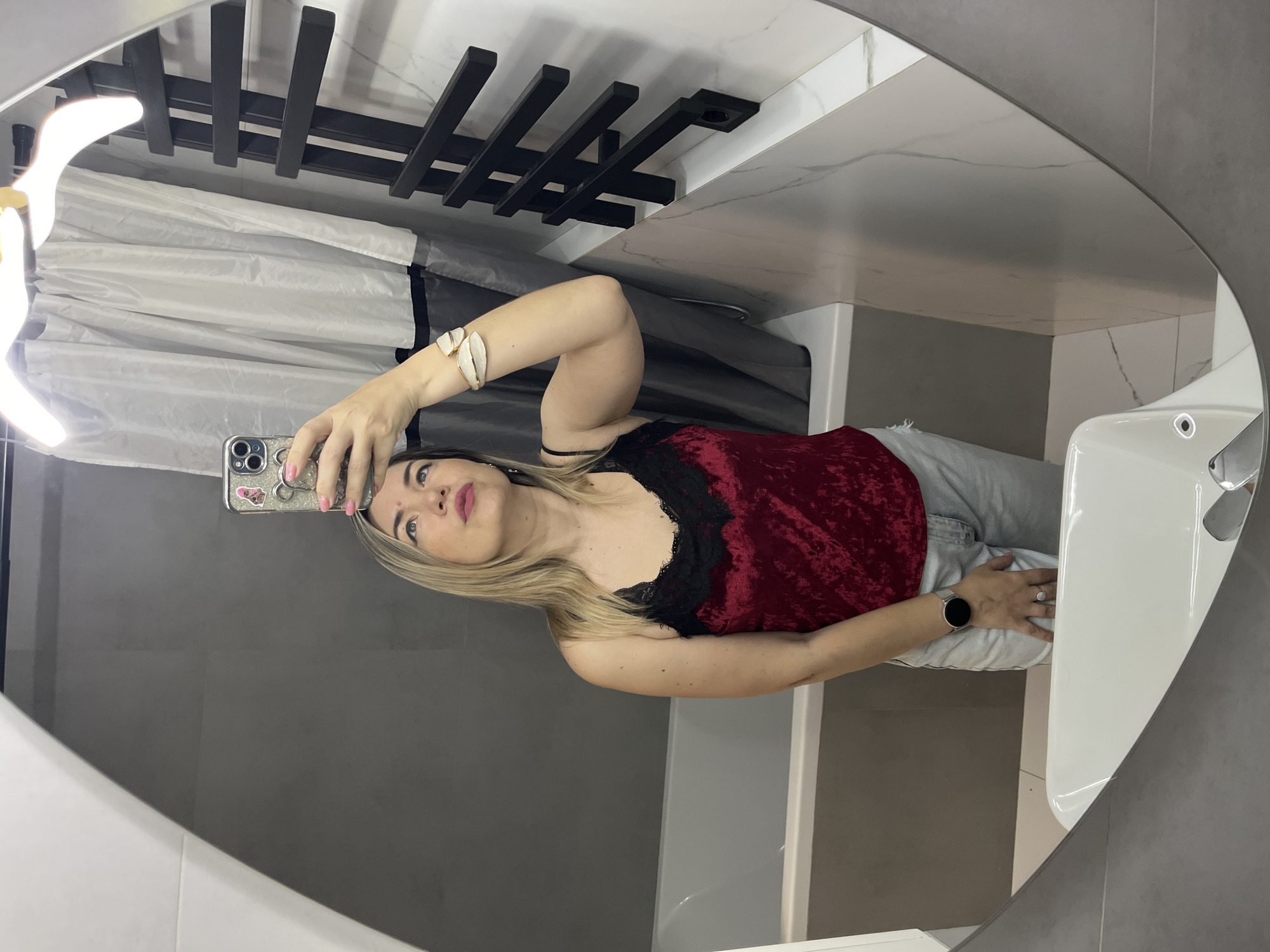 A blonde woman takes a mirror selfie in a bathroom. She is wearing a red velvet camisole with black lace trim, light-wash denim shorts, a silver smartwatch, and a white leaf-shaped bracelet. She is standing in front of a bathtub and a grey tiled wall. A black towel warmer is visible to the right, and a white shower curtain hangs in the background. The bathroom appears to be modern and well-lit, with a large oval mirror reflecting the scene. The image does not provide any information that can be used to determine the location in Sochi, Russia.