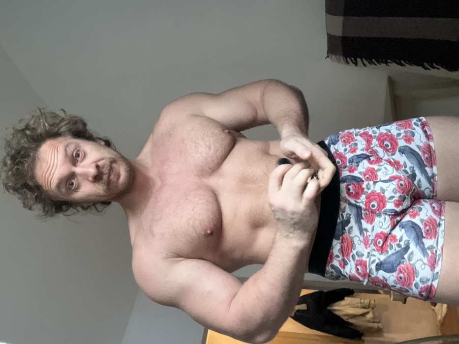 A muscular, shirtless man with curly hair poses for the camera. He wears floral patterned boxer briefs with crows and eye motifs. The background includes a neutral-toned wall and a dark fringed object hanging above what appears to be a wooden surface. The man looks directly at the camera while holding something small between his fingers.