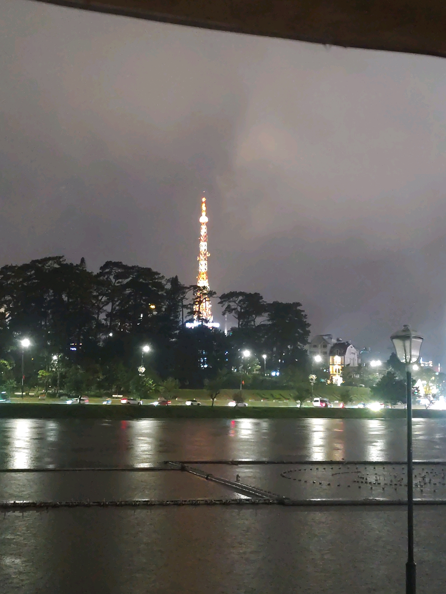 This nighttime, low-light image depicts a panoramic view of Xuan Huong Lake in Da Lat, Vietnam. In the distance, a brightly lit transmission tower stands tall against a cloudy sky, its colorful lights a prominent feature of the cityscape. The lake's surface reflects the lights from the surrounding area and the overcast sky, creating a shimmering effect. Along the lake's edge, a line of trees forms a dark silhouette, punctuated by streetlights and illuminated buildings. A road runs parallel to the lake, with a few vehicles visible, their headlights and taillights creating streaks of light. In the foreground, the lake's surface appears calm, with faint patterns of what may be floating structures or debris visible beneath the water's surface. A lamppost on the right side of the frame adds to the ambient lighting of the scene. The overall atmosphere is subdued and tranquil, characteristic of a quiet evening in Da Lat.