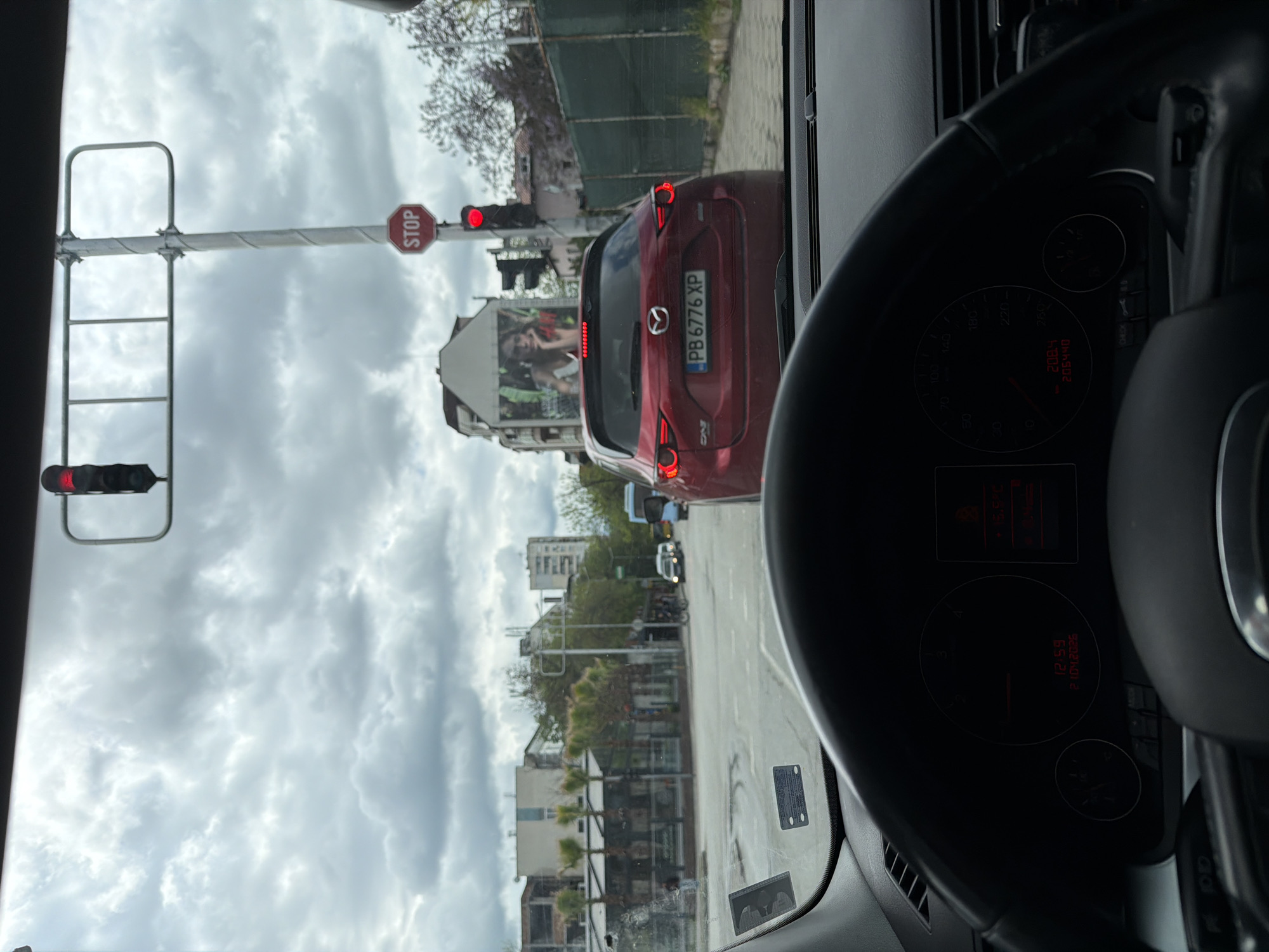 A street-level photograph, taken from the interior of a car, captures a scene at a traffic light under an overcast sky in Plovdiv, Bulgaria. The image is oriented vertically (sideways).
In the immediate foreground, parts of the car's dark dashboard and steering wheel are visible on the right. The instrument cluster shows the car is stationary (speedometer at 0), with a total mileage of 206,440 and a trip meter reading of 208.4. The digital display indicates the time as 12:59, the date as 21/04/2026, and an outside temperature of 15.5°C.
Ahead of the car, a red Mazda CX-5 SUV is stopped at a red traffic light. Its taillights are illuminated. The vehicle's Bulgarian license plate, "PB 6776 XP", is clearly visible, with "PB" signifying Plovdiv, confirming the provided location context.
To the left of the Mazda, a traffic light pole extends across the frame. A red "STOP" sign is attached to the pole, and the traffic lights below it are red, indicating that traffic is halted.
The background reveals a typical urban environment. Buildings of varying heights and architectural styles line the street. A large billboard featuring a person is visible behind the red car. Trees with some foliage and other vehicles can be seen further down the road, with the grey, cloudy sky dominating the upper portion of the frame. The overall impression is of a quiet midday traffic stop in a European city.