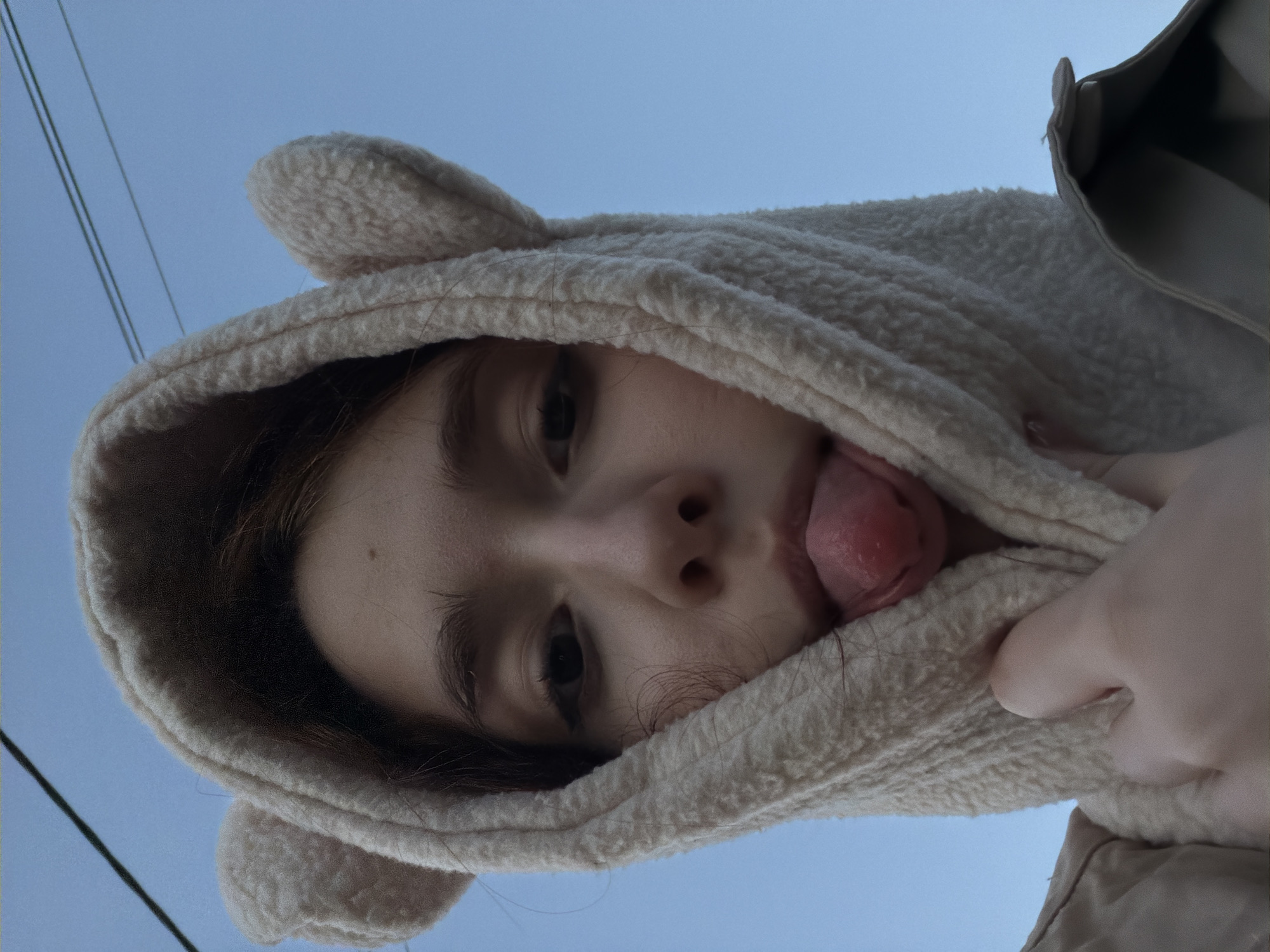 This is a close-up, low-angle selfie of a young person wearing a cream-colored, fuzzy hoodie with bear ears. The person's face is partially obscured by the hood, with only their eyes and mouth visible. They are sticking their tongue out, which is slightly curled. The background is a clear, pale blue sky, suggesting it is daytime. The overall tone is playful and candid. There are a few thin, dark lines visible against the sky, which could be power lines. The subject appears to be outdoors, given the sky and potential power lines. The image captures a moment of lightheartedness, with the subject seemingly posing for a fun selfie.