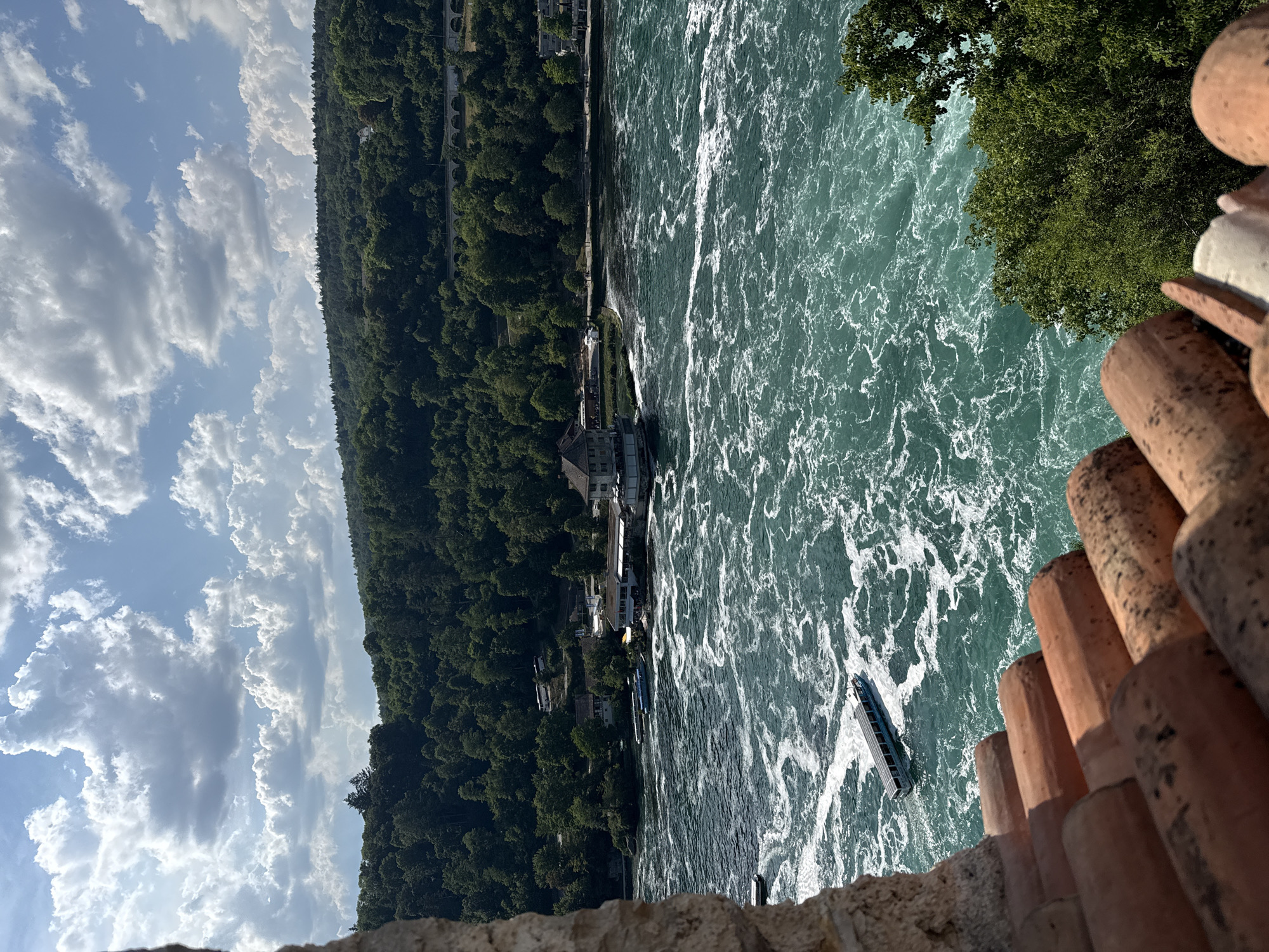 The image depicts a scenic view of the Rhine Falls in Dachsen, Switzerland. The primary subjects are the powerful, turbulent water of the falls, which is a vibrant turquoise color with white frothy waves. A few boats are visible on the water, one with people on board, suggesting a tourist activity. The river is bordered by a lush, green, densely forested hill that slopes down to the water's edge. A few buildings, likely restaurants or visitor facilities, are situated along the riverbank, nestled among the trees. In the foreground, the edge of a stone wall and some reddish-brown, cylindrical objects, possibly roof tiles or decorative elements, frame the view.
The scene is set under a bright sky filled with dramatic, fluffy white clouds against a clear blue backdrop, indicating fair weather. The lighting suggests it is daytime, likely late morning or early afternoon, with sunlight glinting off the water. The overall mood is one of natural grandeur and outdoor leisure. The image is captured from an elevated vantage point, looking down and across the falls. No visible text is present in the image.