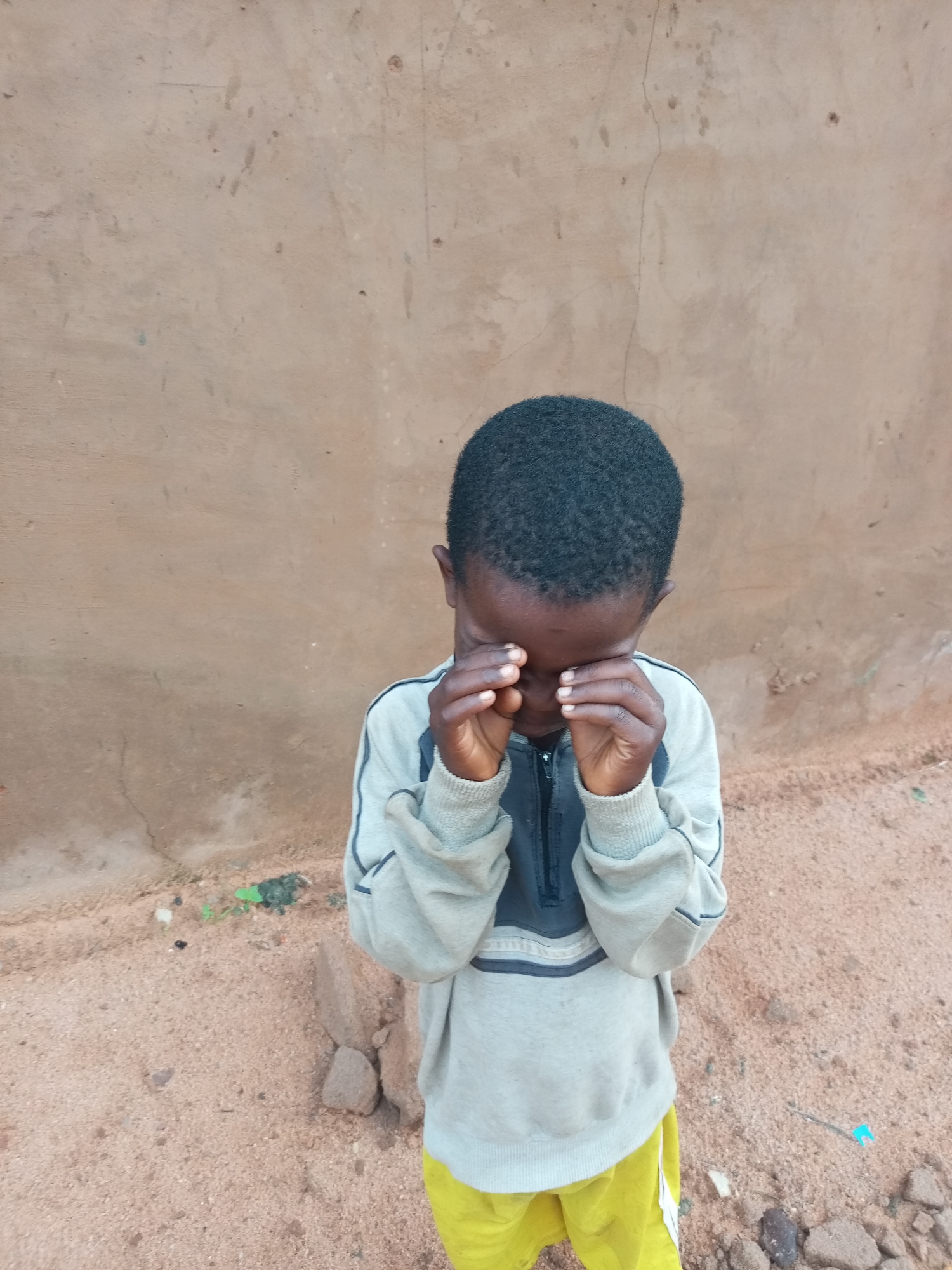A young Black child, appearing to be male, stands with his hands covering his eyes against a textured, light brown wall. The child is wearing a light grey sweatshirt with dark blue and white stripes on the chest and sleeves, and bright yellow shorts with a white stripe. The ground is covered in reddish-brown sand with scattered small stones and debris, including a piece of green plastic. The background wall has visible cracks and some darker marks. The child's posture suggests distress or sadness. The image appears to be taken outdoors in an urban or semi-urban environment. Based on the style of the wall and the ground, this could be a location in the city of Gombe, Nigeria, consistent with typical building materials and ground surfaces found in residential or public areas.