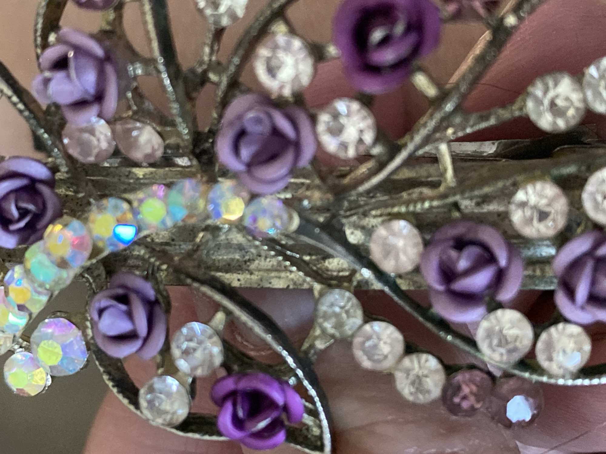 A close-up photograph captures a decorative metallic object adorned with numerous small, intricate purple roses and sparkling rhinestones. The object, possibly a hair clip, brooch, or similar accessory, features a metallic base structured with delicate, swirling branches or filigree designs.
Approximately a dozen small, sculpted roses, varying in shades of purple from deep violet to soft lavender, are attached to the metallic framework. These are complemented by an array of round rhinestones. Some rhinestones are clear and faceted, reflecting white light, while others exhibit a striking iridescent quality, shimmering with vibrant hues of blue, green, yellow, and pink. The metallic base itself appears to have an aged or antiqued finish, showing tones of muted silver and gold, adding to its ornate appearance.
The item is held against a soft, out-of-focus background that reveals a blurred human hand or finger with a reddish-brown skin tone, suggesting the object is being displayed or examined. The foreground and central elements of the decorative piece are in sharp focus, highlighting the details of the roses and the sparkle of the rhinestones.
No specific activity is depicted beyond the presentation of the object. There is no visible text, and the time of day, weather conditions, or any specific emotions cannot be determined from the image. While the location context is Novokuznetsk, Russia, no visual cues within the photograph itself indicate this.