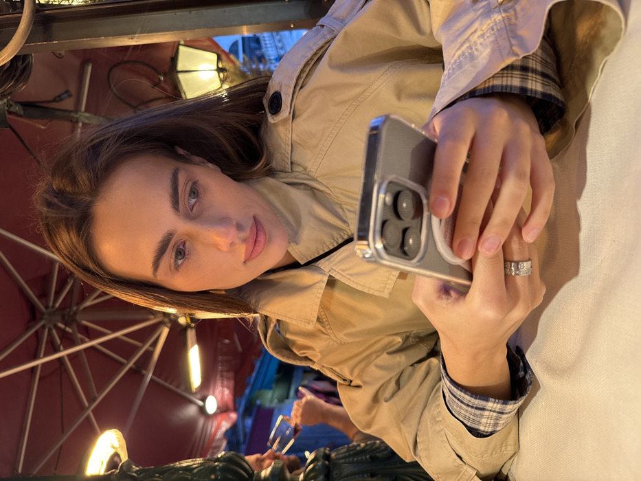 The image depicts a female individual seated at an outdoor cafe table in Venice, Italy, during evening hours. The woman, appearing to be in her late 20s to early 30s with long brown hair, wears a tan trench coat over a plaid shirt. She is focused on a silver-colored smartphone, likely an iPhone Pro model with a clear case and a white circular MagSafe accessory, held in both hands. A decorative ring with etched letters and symbols is visible on her right ring finger. The table is covered with a white tablecloth.
In the blurred background to the left, a male figure is partially visible, raising a wine glass containing red liquid towards his mouth. An ornate, dark column or pillar is positioned next to him. Overhead, a red fabric awning or large umbrella structure, supported by metal ribs, extends across the scene. An orange-glowing outdoor heater is attached to the overhead structure. To the right, a lit, decorative gas lantern-style fixture illuminates the light-colored, classical Venetian architecture of a distant building, featuring arches and columns, characteristic of structures found in prominent areas like Piazza San Marco.
