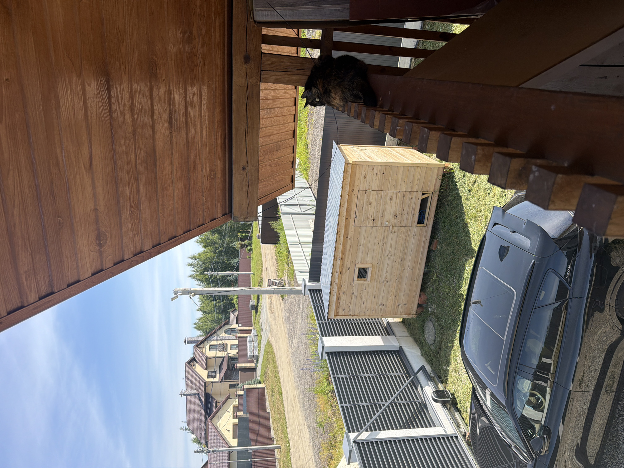 The image depicts a sunny outdoor scene, likely in Malinovka, Russia, showcasing a residential area. In the foreground, a dark-colored car with a panoramic sunroof is parked on a gravel surface. To the left, a modern gray fence with vertical slats borders the property. Adjacent to the fence is a small, unfinished wooden structure with a corrugated metal roof and a single small window. In the background, two multi-story houses with brown roofs and light-colored facades are visible. A gravel road leads between the houses. Lush green trees form a backdrop behind the houses, suggesting a suburban or rural setting. A cat with dark, tortoiseshell fur is perched on a wooden railing in the mid-ground, looking away from the camera. The sky is a clear blue with a few wispy white clouds. The overall impression is one of a tranquil summer day in a developing neighborhood.