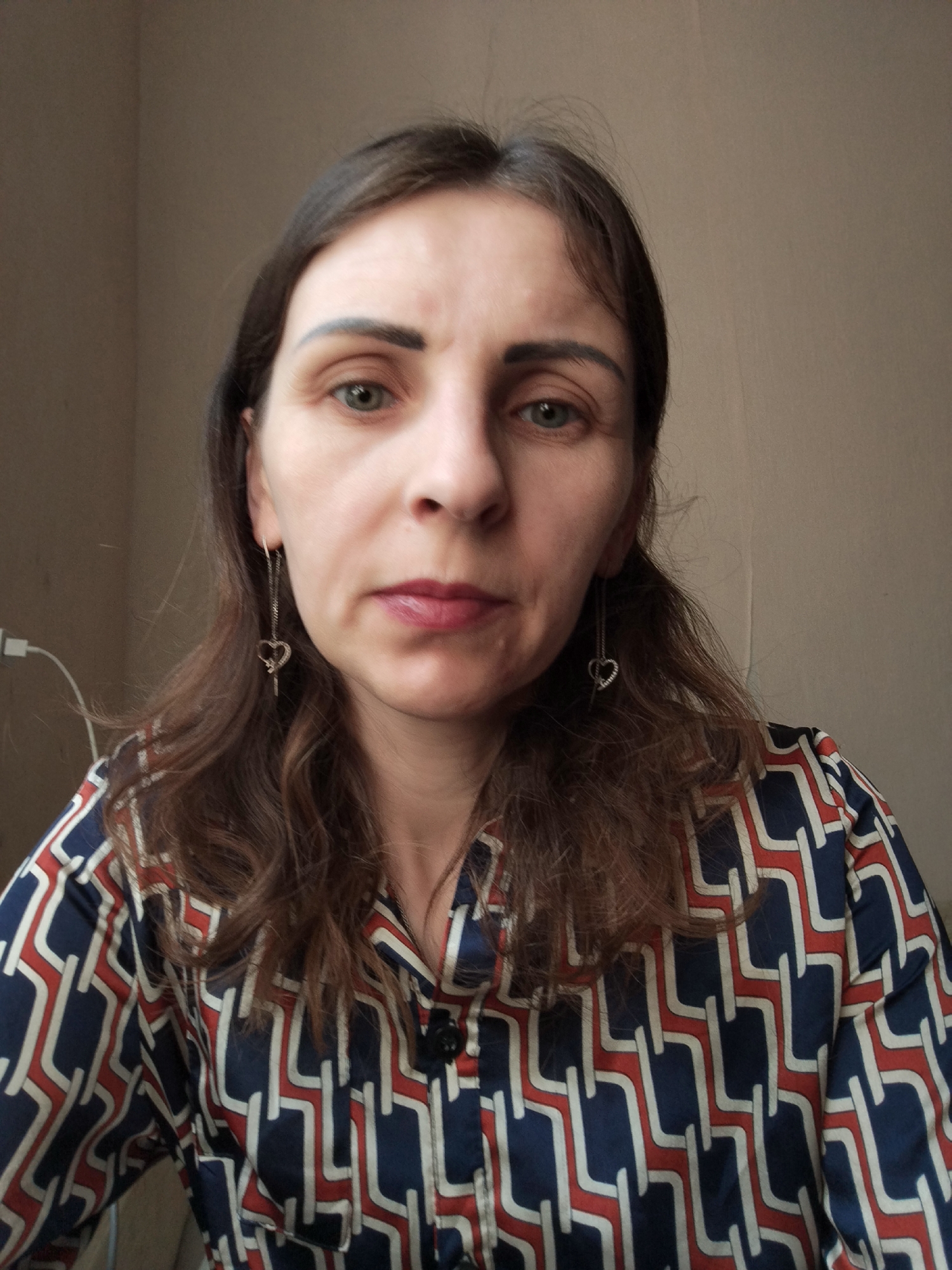 The image shows a medium shot of a woman with long, wavy brown hair and light skin. She is looking directly at the camera with a neutral expression. She is wearing a patterned blouse with dark blue, red, and white geometric shapes. She has on dangling earrings with a heart shape at the end. The background is a plain, light brown wall with some electrical outlets visible on the left side. The scene appears to be indoors, possibly in a home or office setting. No specific location in Vinnytsia, Ukraine can be identified from the image alone.