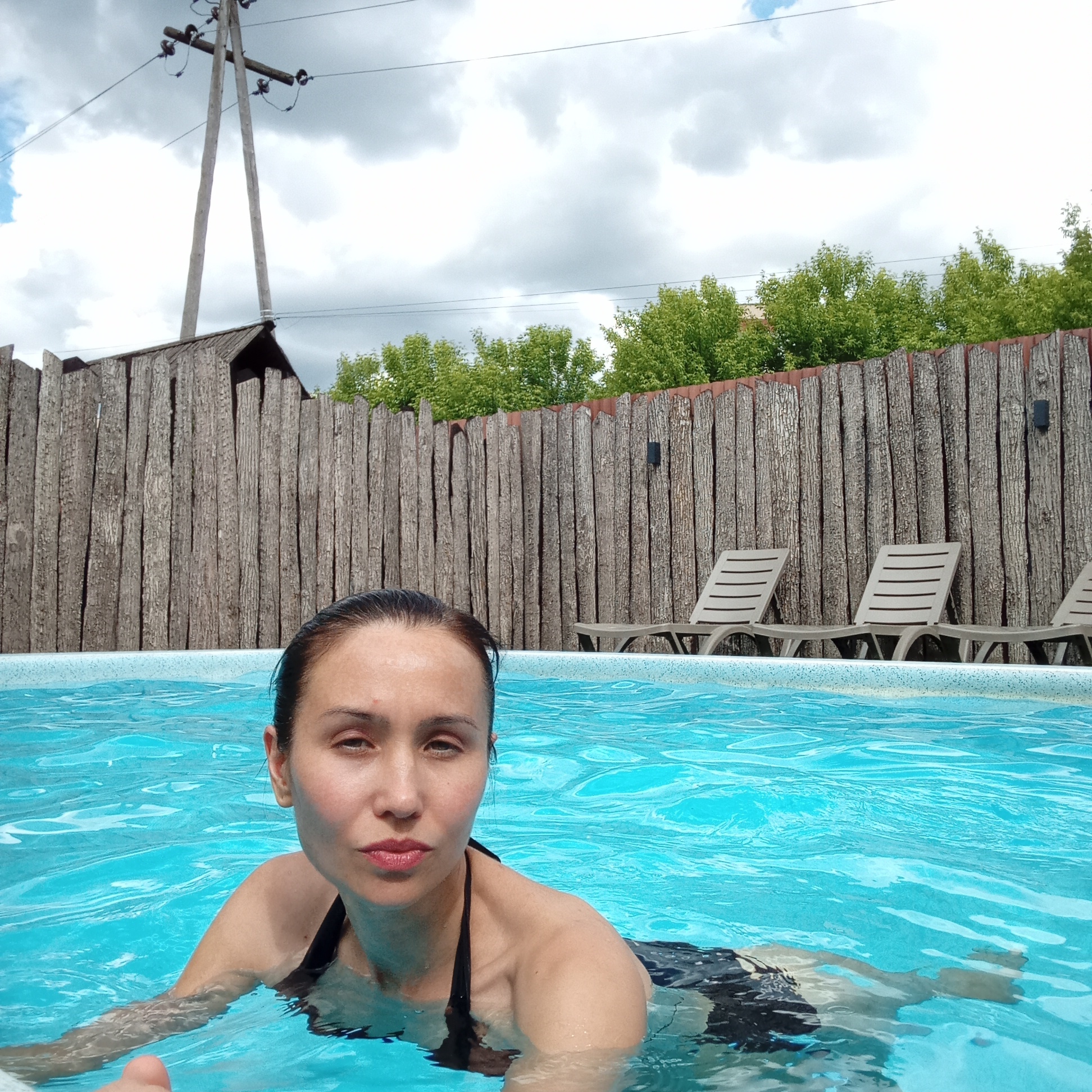A woman is in a swimming pool. Behind her is a fence made of vertical wooden planks and a row of tan plastic lounge chairs. Behind the fence are trees. A utility pole is in the background. The location is outdoors and appears to be in Izhevsk, Russia.