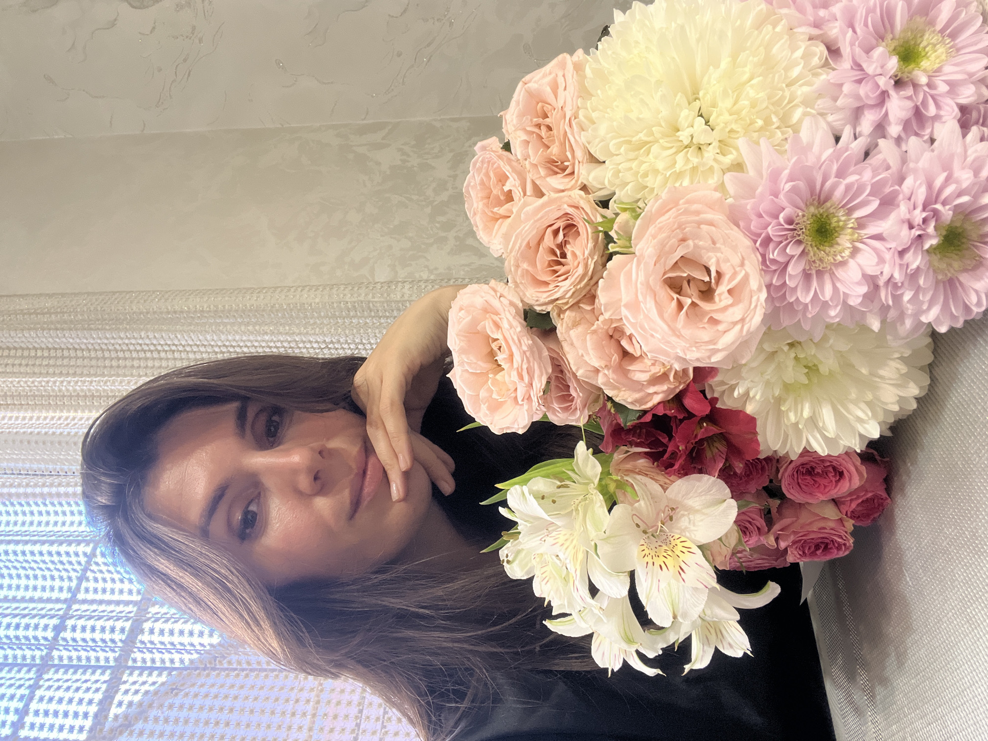 The image captures a serene indoor portrait of a woman holding a voluminous bouquet of flowers. The woman, with medium-length brown hair and dark eyes, is positioned on the left, looking directly at the camera with a gentle, soft smile. Her left hand is delicately placed near her chin. She appears to be wearing dark clothing.
The large bouquet, occupying the right side of the frame, is a vibrant mix of various flowers and colors. Dominant are pale pink garden roses with numerous ruffled petals, alongside large white or cream chrysanthemums. Clusters of light purple chrysanthemums with distinct green centers add another pastel hue. White alstroemeria (Peruvian lilies) with delicate yellow and reddish-brown markings are also visible, complementing a few smaller, deep red or burgundy roses.
The background behind the woman features a light-colored, textured fabric, possibly blinds or curtains, with a subtle grid-like pattern, suggesting a window and natural light source. Above this and behind the flowers, a light beige or grey wall with a faint, abstract, and possibly glossy pattern can be seen. The overall lighting is soft and even, typical of an indoor setting. The atmosphere is calm and appreciative. No visible text is present in the image. The location context provided is Kaspiysk, Russia.