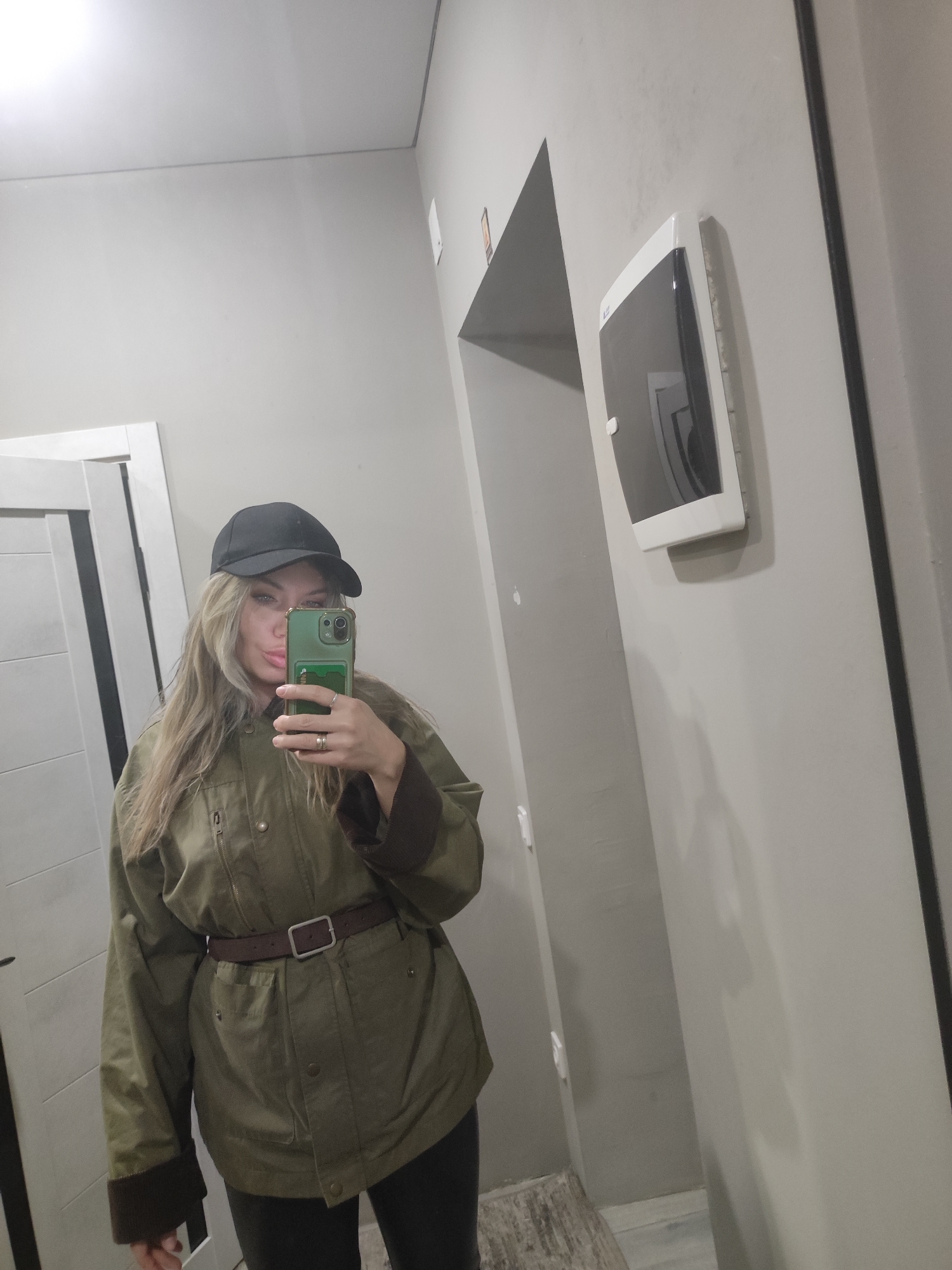 A woman in an olive green jacket with a brown belt and a black baseball cap is taking a selfie in what appears to be a hallway. She is holding a green phone. The hallway has gray walls, a white door frame, a mirror, and a white object mounted on the wall. There are no indications that this image was taken in the city of Votkinsk, Russia.