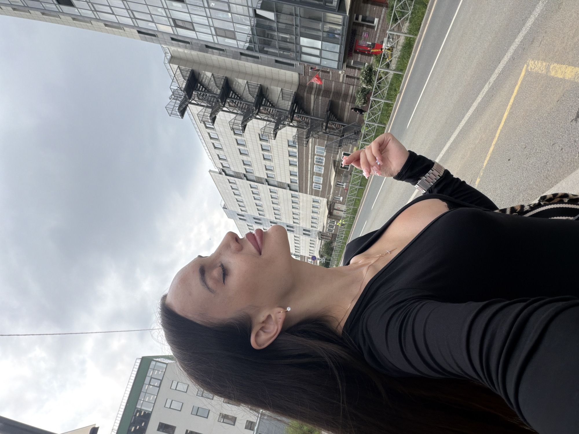 A woman with long, dark hair, wearing a black, low-cut, long-sleeved top and a cross necklace, stands on a sidewalk next to a street. She is looking to her right with her eyes closed and her head tilted back slightly, with a serene expression. She is wearing a silver earring in her left ear and a silver watch on her left wrist. Her right hand is raised, with her fingers curled and painted with pink and white acrylic nails. In the background, there are modern, multi-story buildings with glass facades and balconies, some with external metal staircases. A street with a yellow dividing line is visible in the foreground. The sky is overcast and cloudy. The scene appears to be an urban environment in Saint Petersburg, Russia.