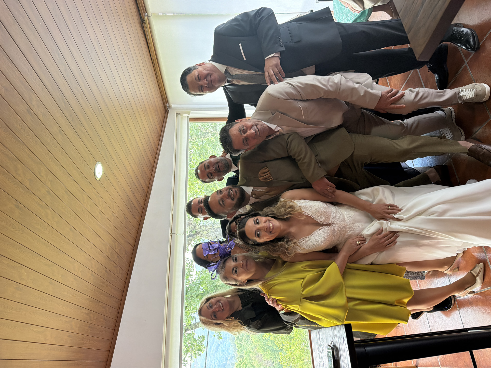 A group of eight people are stacked on top of one another. Starting from the bottom, a blonde-haired woman in a yellow dress lies flat. Above her is a woman wearing a purple fascinator, followed by a bride in a white wedding dress. Above her are four men in suits, in varying shades of tan and brown, and finally a man in a dark suit. All the people in the photograph are smiling and appear to be having fun. Behind the group is a window overlooking trees. The interior of the building has a wooden panelled wall. The location is Vila Real, Portugal.