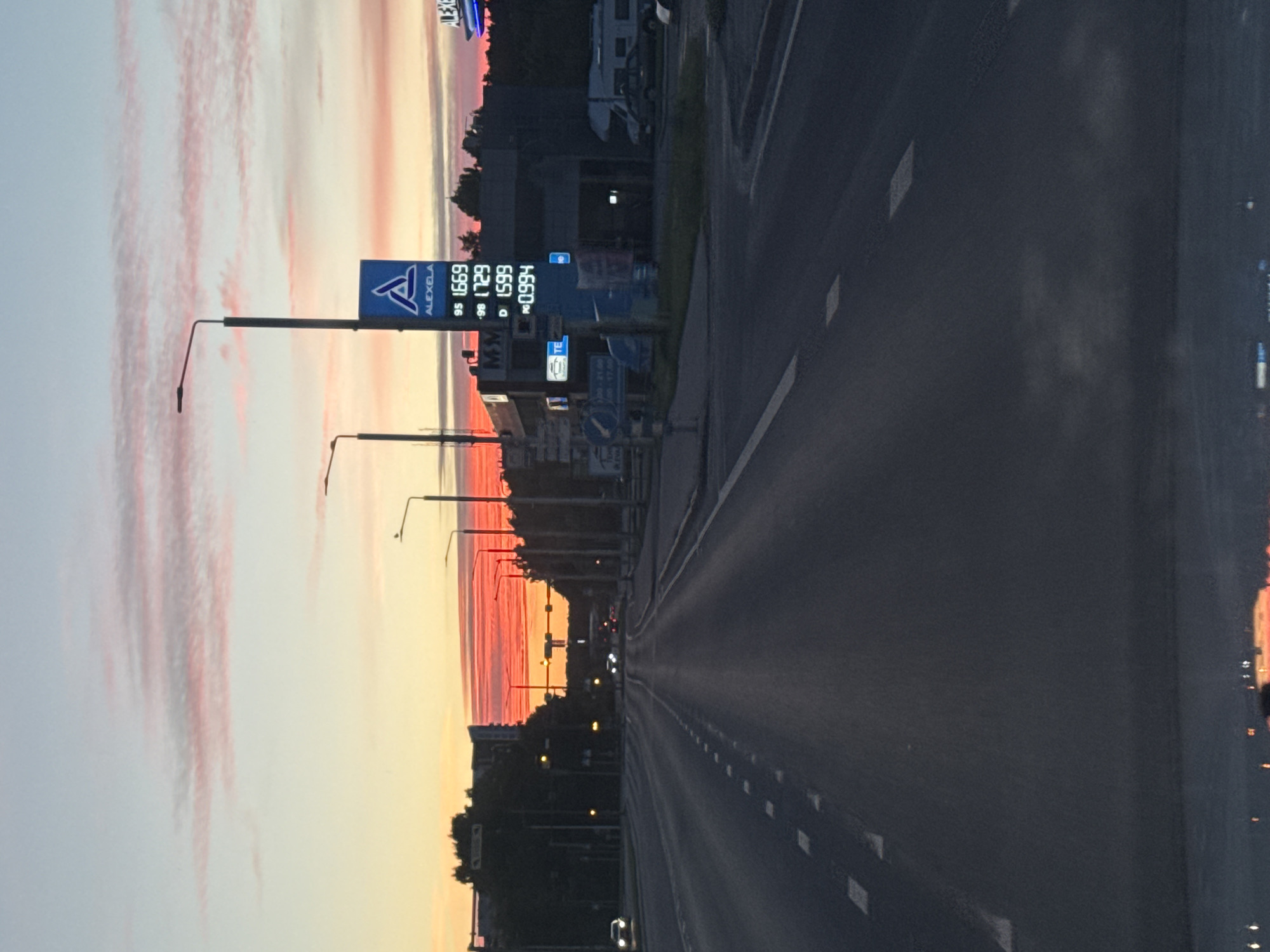 A low-angle, full-shot perspective shows a dark asphalt road stretching towards a vibrant sunset over Tallinn, Estonia. The sky is a gradient of soft pink and pale blue, with streaks of orange and red along the horizon. Streetlights and buildings are silhouetted against the colorful sky. A prominent blue sign for "ALEXELA" displays fuel prices, with "95 1.669", "98 1.729", and "D 1.599" visible, along with another price of "0.994" likely for something else. A parked RV and a dark car are visible to the right of the gas station. Several cars are on the road, their headlights illuminated. The scene appears to be captured from inside a vehicle, as indicated by the dashboard visible at the bottom of the frame. The overall impression is one of urban twilight, with a focus on the road and the sky.