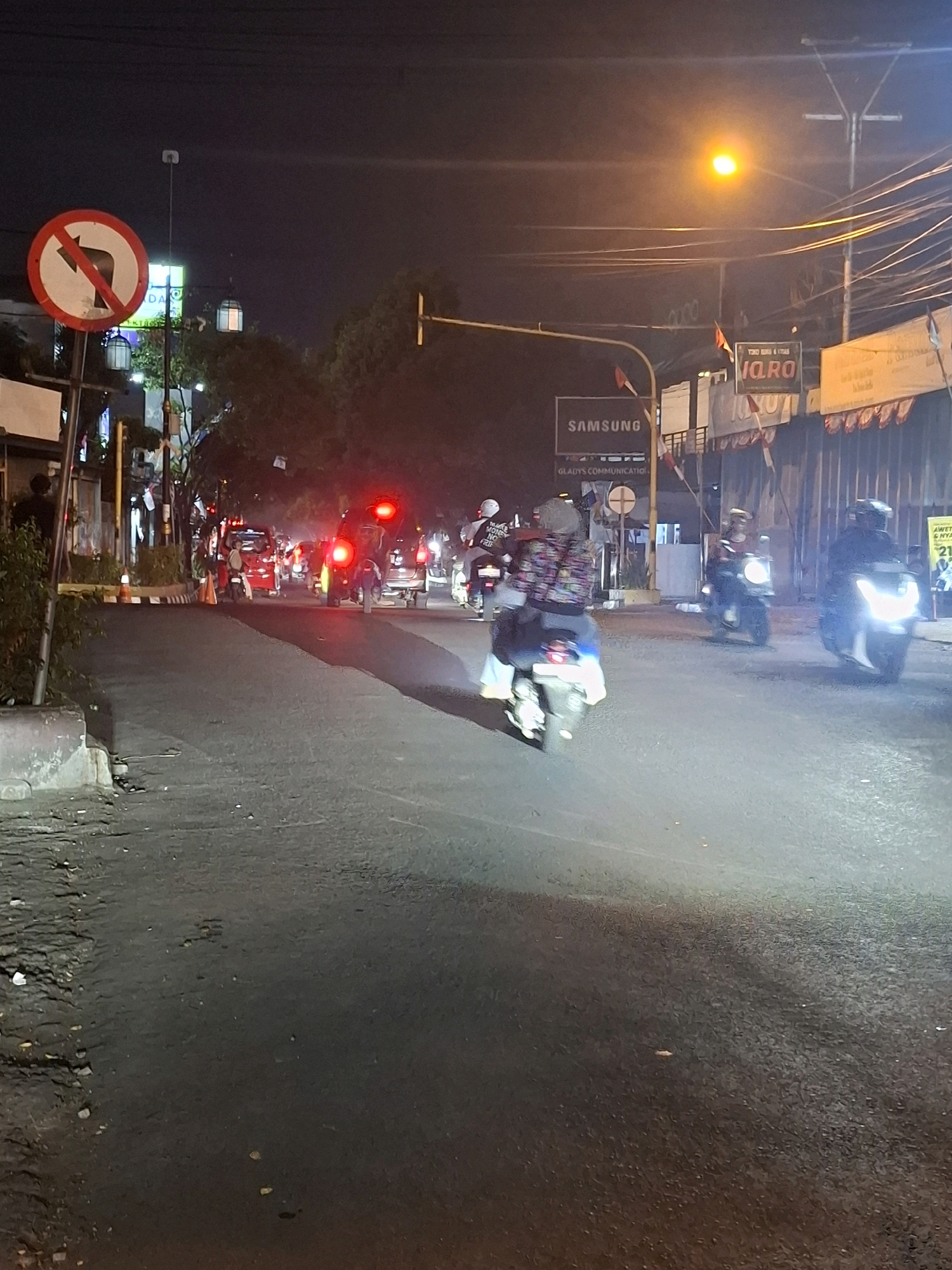 The image depicts a street scene at night in Cianjur, Indonesia, likely during a period of moderate traffic. A traffic sign prohibiting right turns is visible on the left. Multiple motorcycles with riders are present on the road. In the foreground, a motorcycle is seen from the rear, with its taillights illuminated. Further down the street, other motorcycles and a few cars are visible, some with their headlights and taillights on. Buildings with illuminated signs, including "SAMSUNG" and "VARA," line the right side of the street, suggesting a commercial area. Streetlights and overhead wires contribute to the overall illumination of the scene. The road surface appears to be asphalt.