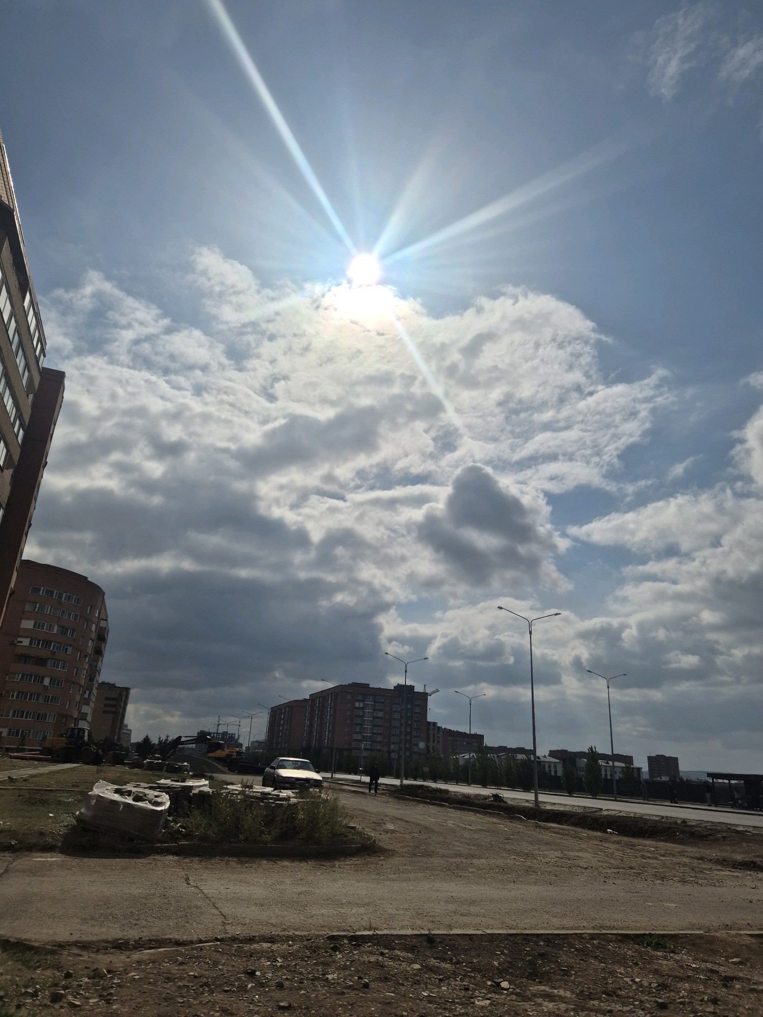 The image shows a street scene in Kokshetau, Kazakhstan, on a bright, cloudy day. The sun is shining brightly in the upper center of the frame, creating a sunburst effect. In the foreground, there is a dirt road with some construction materials, including wrapped bundles of what appear to be building supplies and some loose bricks. To the left, a tall, modern apartment building with a brick facade dominates the left side of the image. In the middle ground, across a paved road with streetlights, are several more apartment buildings. A white car is parked on the side of the road, and two individuals are walking in the distance. The background shows more buildings and trees, with the sky filled with a mix of bright white and dark gray clouds. The overall impression is of an urban environment, possibly undergoing some construction or development.