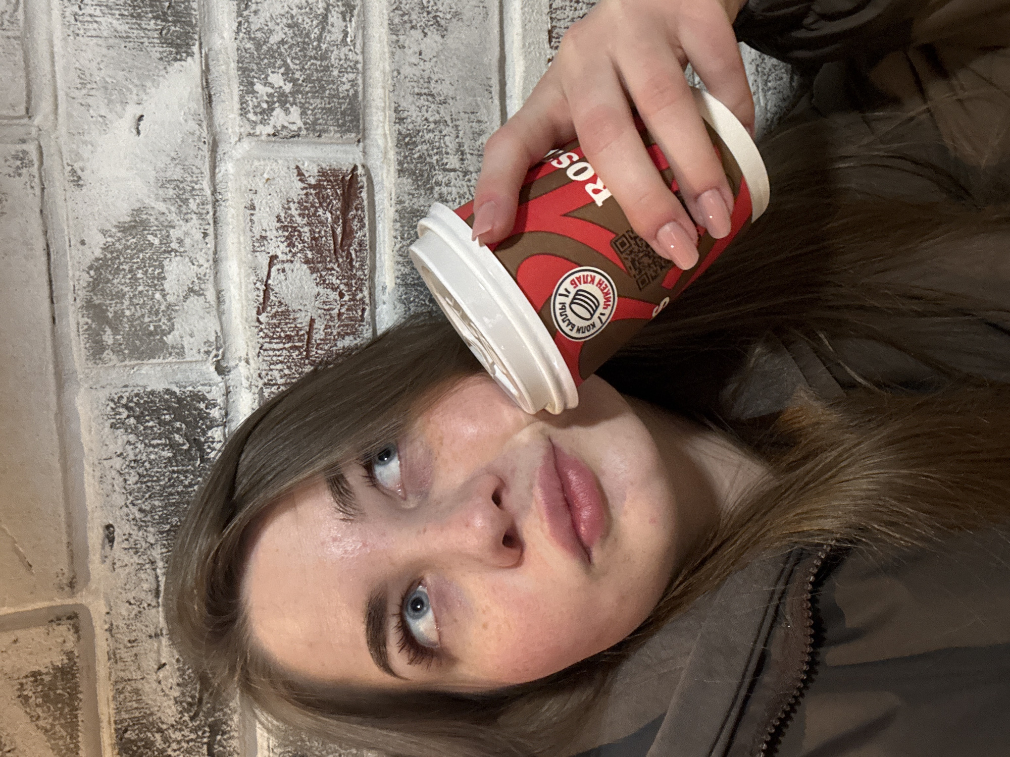 A young woman is depicted in a close-up shot, looking upward and slightly to her right. She has light brown hair, striking blue eyes, and fair skin with subtle freckles. Her lips are naturally full and have a pink hue. She is wearing a dark, possibly brown or grey, jacket, with a glimpse of what appears to be a fur collar or hood around her neck.
An activity is taking place where a disposable coffee cup is casually balanced on her chin, held by her right hand. Her fingernails are neatly manicured with a light pink polish.
The setting appears to be indoors, against a white-painted brick wall that shows signs of wear, with some of the underlying reddish-brown brick visible. The lighting is soft and even, typical of an indoor environment. Her expression is neutral and contemplative.
Visible text on the coffee cup includes a partial brand name "ROS" (possibly part of "ROSTIK'S"), a QR code, and a circular logo featuring stacked cups. Around the logo, Cyrillic text reads "КОПИ БАЛЛЫ" (Collect points) and "ВЫЙГРАЙ КУБОК" (Win a cup). The image provides a glimpse into a moment in Chaikovsky, Russia.