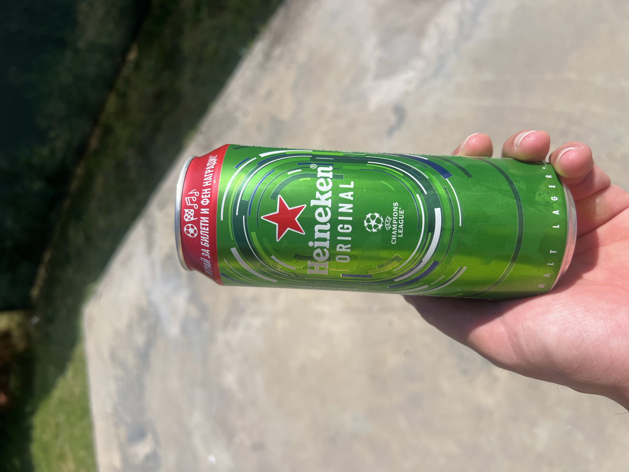 A person holds a green Heineken Original can in their right hand. The can features the Heineken logo with a red star and the Champions League logo. The text on the can is in English and another language. The background consists of concrete and green vegetation, suggesting an outdoor environment. The image was probably taken during the day. The specific location in Herakovo, Bulgaria is not determinable from the image alone.