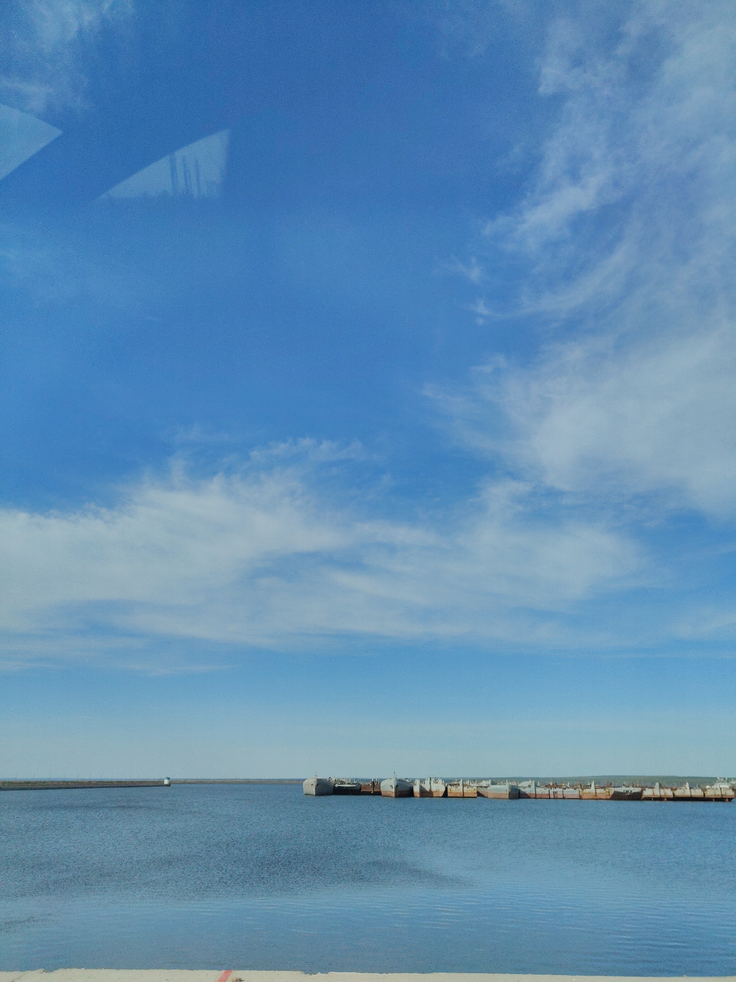 The image depicts a body of water with a pier in the distance and a series of docked ships or barges towards the right. The sky is blue with scattered white clouds. A faint reflection or glare is visible in the upper left corner. The location is Unknown, Unknown.