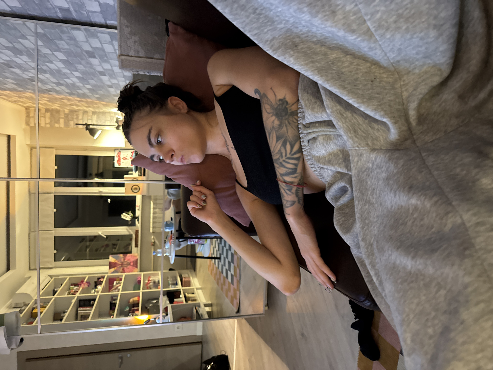A young woman with dark hair tied in a bun is sitting on a couch, looking to the left with a neutral expression. She is wearing a black tank top and grey sweatpants. Her left arm is resting on a pillow, and she has a tattoo on her left arm. The couch appears to be in a living room, with a large mirror reflecting shelves filled with items, a table with chairs, and windows. The room has brick-patterned walls and wooden flooring. The overall lighting suggests it is evening. There are no specific indicators of the city being Moscow, Russia.