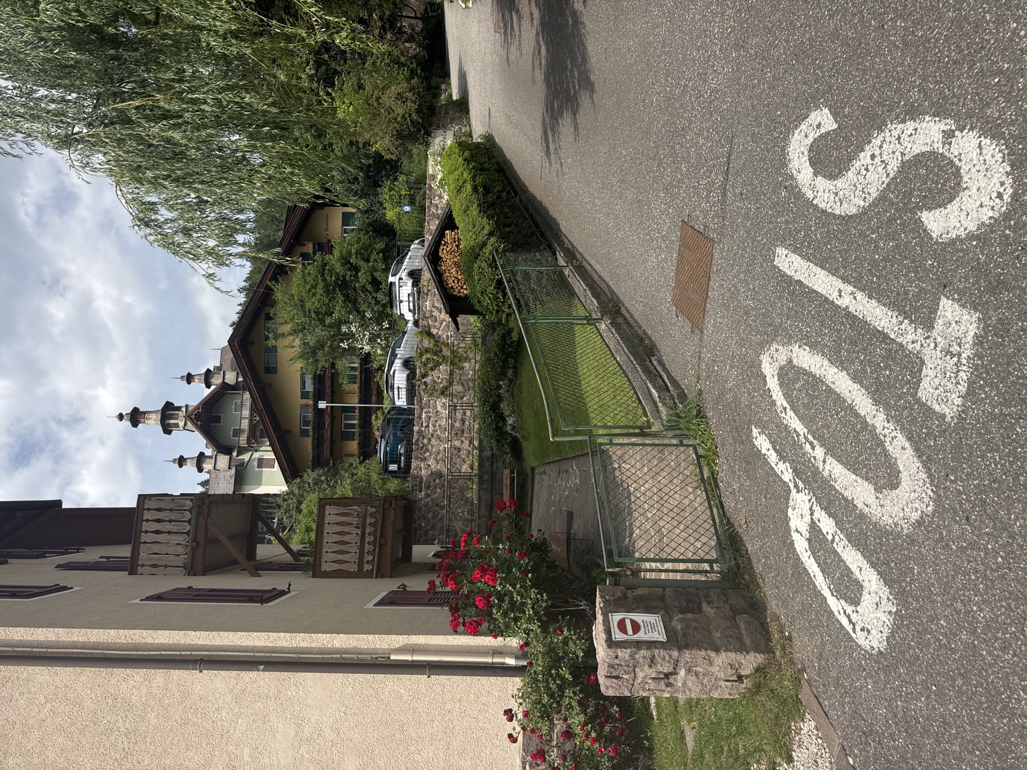 The image depicts a residential street in Urtijëi - St. Ulrich in Gröden - Ortisei, Italy. The scene is a view from a slight downward angle, looking across a paved road with a white "STOP" sign painted on it. The road is bordered by a green metal fence, beyond which is a well-maintained lawn and a stone wall with a sign indicating a prohibition. Adjacent to the lawn is a small wooden structure filled with firewood, nestled behind a dense green hedge.
Further into the background, several houses with traditional alpine architecture are visible. One house is a light yellow color with dark wooden trim and shutters, featuring decorative balconies. Another building, taller and with a more ornate roofline, has multiple spires or towers. Several cars are parked along the side of the road. A lush green tree with long, drooping branches dominates the upper left of the frame. A vibrant bush with bright red roses is in the foreground, near the bottom right.
The lighting suggests it is daytime, possibly late morning or early afternoon, with some clouds in the sky. The overall impression is one of a peaceful, picturesque European village. The visible text includes the word "STOP" on the road and text on a white sign with a red circle and line, indicating a prohibition.