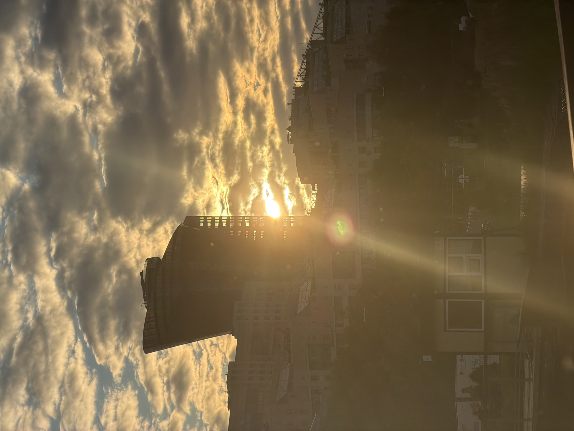 This is a vertically oriented, wide-angle photograph capturing a cityscape bathed in the warm light of sunrise or sunset. The sun, a bright, blinding orb, is positioned centrally, peeking through dramatic, wispy clouds that are illuminated in hues of gold and white. Sunlight beams cut through the atmosphere, creating a lens flare with a green tint near the center of the image.
Dominating the scene are tall buildings, their architectural details partially obscured by the intense light and the angle of the shot. One prominent building on the left has a curved, stepped facade and appears to be under construction or renovation, with scaffolding visible. Another building on the right is a lighter-colored, multi-story structure with numerous windows. Trees and foliage are visible at the base of the buildings, adding a touch of nature to the urban environment. The overall impression is one of a dynamic urban landscape experiencing a moment of striking natural beauty, with the sun's rays dramatically interacting with the clouds and man-made structures. The time of day is clearly either dawn or dusk, indicated by the low angle and warm color of the sun, and the dramatic cloud formations.