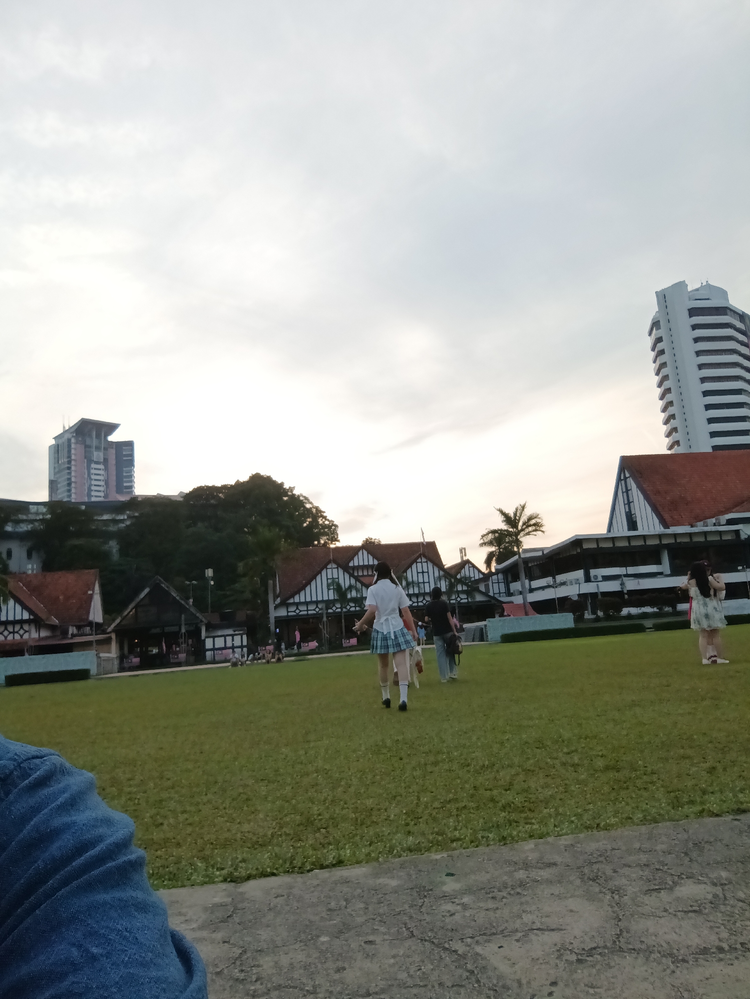 The image captures a scene in Kuala Lumpur, Malaysia, featuring a wide grassy field in the foreground, with a group of people walking across it. Several low-rise buildings with red tiled roofs and distinctive white trim are visible in the middle ground, possibly part of a heritage site or a university campus. In the background, a tall, modern high-rise building with a white facade stands out against the overcast sky. A few trees, including a palm tree, are scattered across the landscape, and the overall atmosphere suggests it is late afternoon or early evening due to the soft light. The perspective is from ground level, looking towards the buildings and the sky.