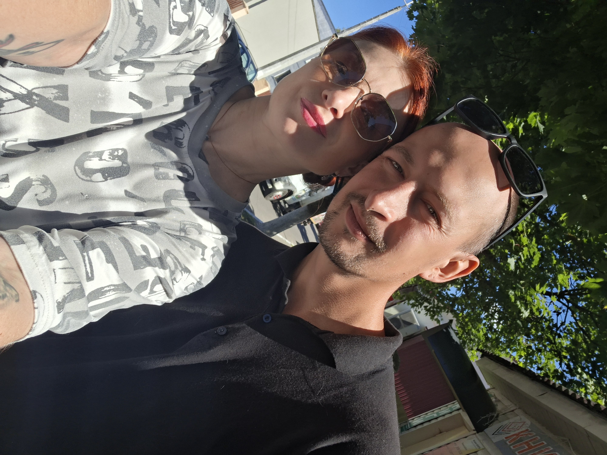 A selfie taken outdoors in Myadzyel, Belarus, features a man and a woman. The man, on the right, has sunglasses perched on his head and is wearing a black polo shirt. The woman, on the left, wears sunglasses and a gray t-shirt with a white and gray pattern of numbers and letters. They are standing close together, with the man's arm possibly around the woman. In the background, a tree with green leaves is visible, along with a building with a sign and a white truck. The sky is blue, suggesting a sunny day.