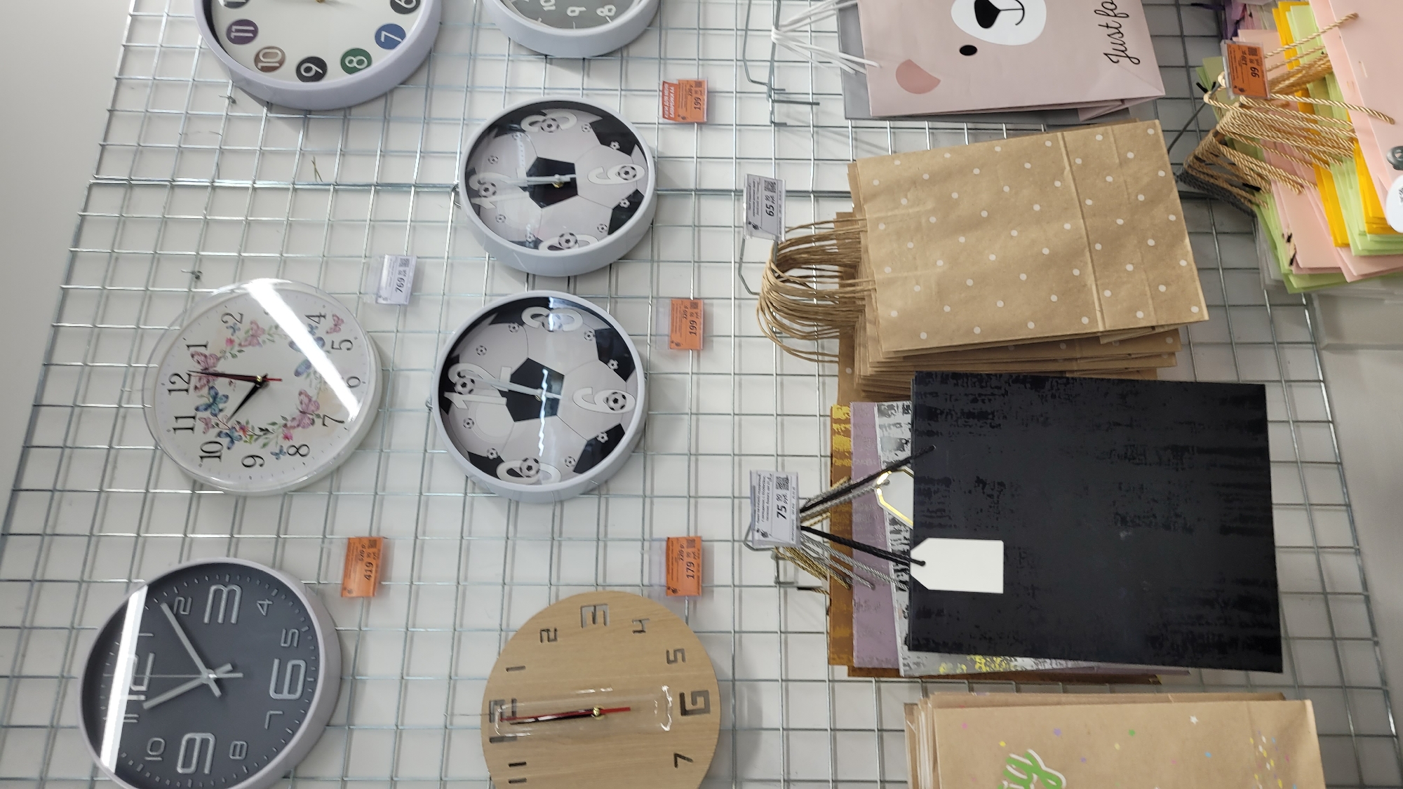 Captured from a high-angle perspective, the image showcases an array of wall clocks and gift bags affixed to a white wire grid. The clocks vary in style and color, with some featuring conventional numerals and hands, while others showcase unique designs like soccer balls or butterflies. The gift bags come in an assortment of sizes and colors, some adorned with minimalist designs like polka dots or bear faces. Small price tags are visible near several items, suggesting they are for sale. The overall composition indicates a retail environment, possibly a shop or market stall.