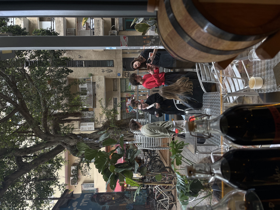The scene depicts an urban street environment in Tel-Aviv, Israel, characteristic of areas such as the Carmel Market or Nachalat Binyamin.
In the immediate foreground, out of focus, two clear glass bottles containing a dark liquid are visible, one with a swing-top closure. To the far right, a partial view of a wooden barrel with metal bands is present. A small white ceramic cup and a clear glass rest on a textured, silver-colored metal outdoor café table. A person with light brown hair, wearing a dark jacket, is seated at a similar table, facing away from the camera.
The midground features six individuals gathered on a paved street area, presumably an outdoor café seating zone. A man with light hair and a beard, dressed in a light green long-sleeved shirt and light pants, stands holding a dark bag. To his right, a woman with glasses and dark hair, wearing a black coat, looks down at a mobile phone while holding a white cup. Next to her, a man with dark curly hair, wearing a red long-sleeved shirt and dark pants, observes his mobile phone. Another woman with dark hair, in a black t-shirt, stands beside him, also looking at a mobile phone and holding a white cup. An additional person is partially obscured behind the man in the green shirt. Behind the standing group, several silver-colored café chairs are stacked. Various potted plants with large green leaves are interspersed among the seating arrangements.
The background reveals a multi-story urban landscape under what appears to be an overcast sky. A large, mature tree with extensive branches and visible aerial roots dominates the center-left. To the left, a building features a blue mural with a stylized portrait and Hebrew text, including "תודה" (Toda - Thank you) and "שוק" (Shuk - Market). Numerous bicycles are parked along the street to the left. The central background features a beige residential or mixed-use building with multiple windows, balconies, external air conditioning units, and white shutters. Graffiti, including the Hebrew word "אורח" (Ore'ach - Guest), is visible on the building walls. A street number "37" is affixed to the building wall on the right.