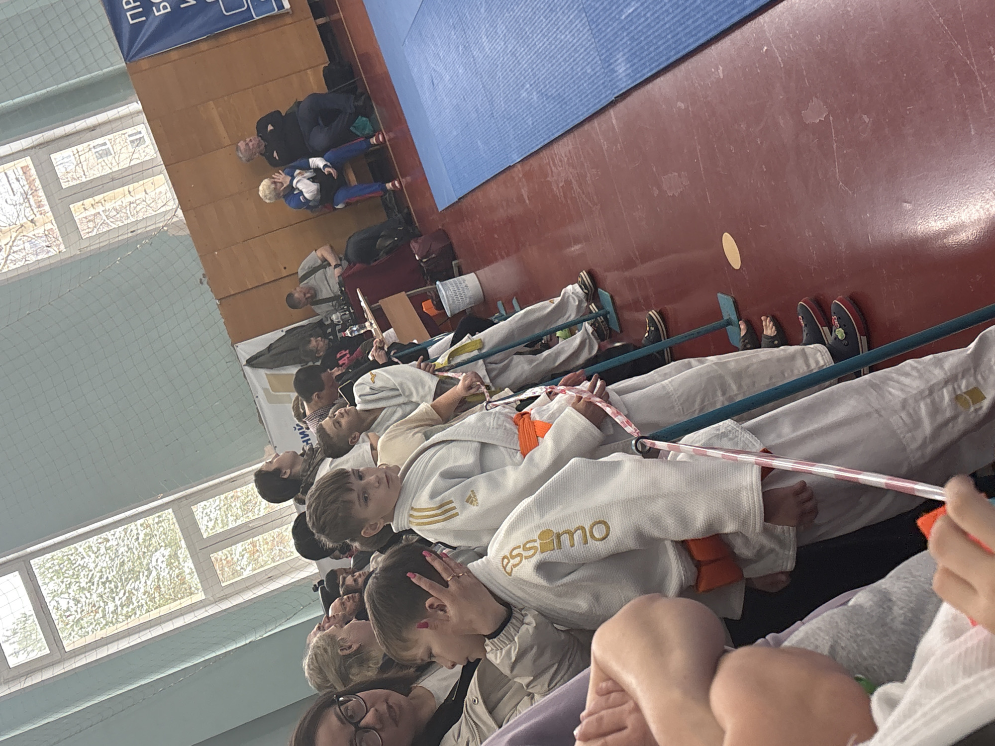 In Chaikovsky, Russia, a group of individuals wearing judo uniforms are seated indoors, possibly waiting for an event. A blue judo mat is visible in the background, along with windows allowing natural light into the room. Some adults are also seated further back, observing the scene.