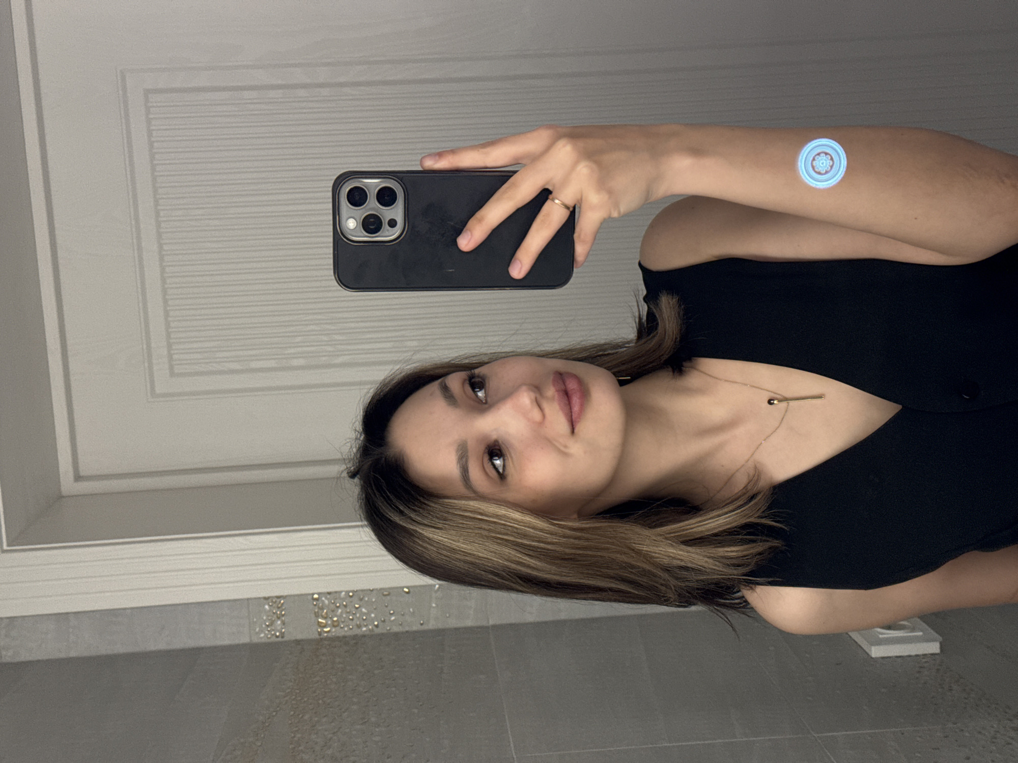 A young woman is taking a selfie in what appears to be a bathroom. She is holding a dark-colored smartphone with a multi-lens camera. She is wearing a black top and a gold necklace. There is a blue, round object on her arm. The background includes a white door with panel detailing and tiled walls with decorative elements. The location is Unknown, Unknown.