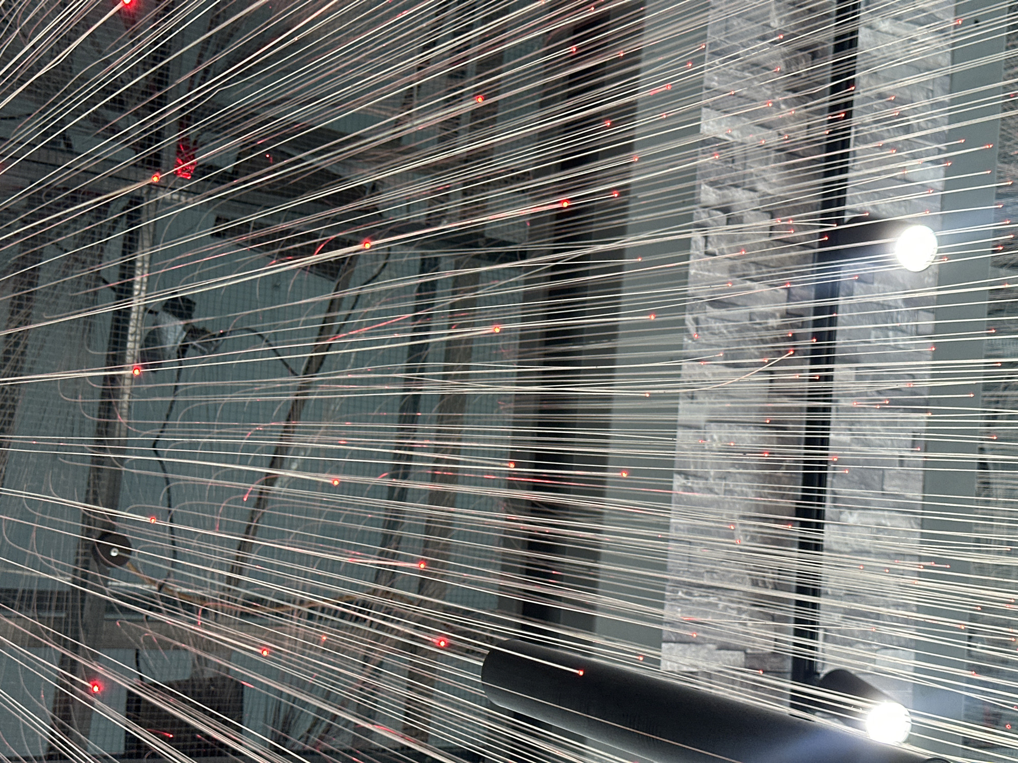 The image depicts an artistic installation featuring a dense array of parallel, illuminated strands emanating from a focal point. The strands appear white or silver with small, randomly distributed red points of light. In the background, a wall constructed of gray bricks can be seen. The overall aesthetic is modern and potentially abstract. The presence of the brick wall as a backdrop might place the installation outdoors or in a gallery-like setting. The presence of the tubular object could either be a part of the installation or it is the result of the viewer's close perspective. The image is from Milan, Italy.
