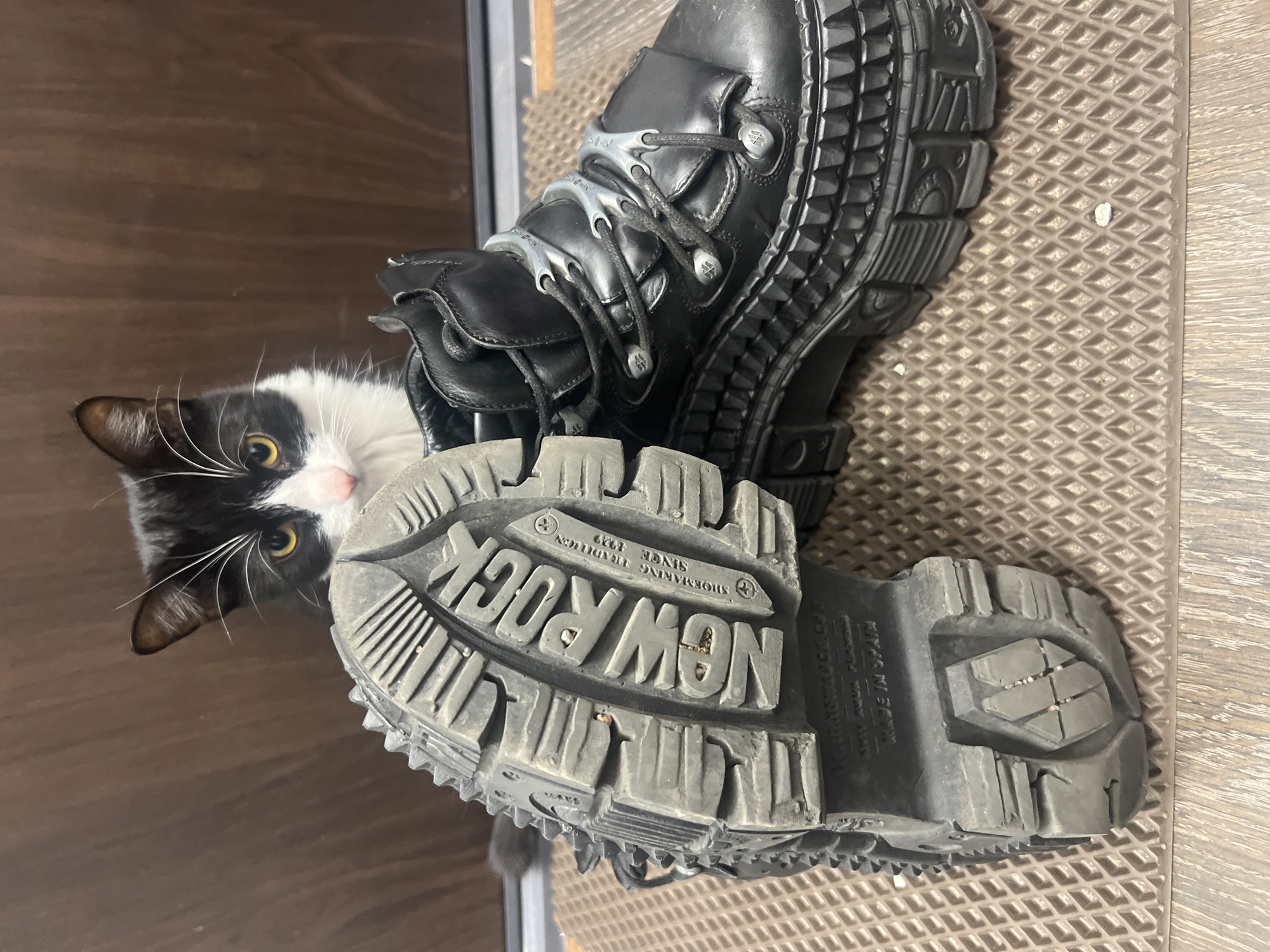 This image depicts an indoor scene in the city of Unknown, Unknown, featuring a tuxedo cat peeking from behind two black platform boots. The cat, exhibiting black fur with prominent white markings on its face, chin, and chest, has distinct yellow eyes and long white whiskers. It is positioned centrally in the frame, with its head slightly raised above the sole of the boot on the left.
Two black, heavily-soled platform boots are present. The boot on the left is oriented on its side, revealing its grey rubber sole, which bears the embossed brand name "NEW ROCK" in large lettering, along with smaller text indicating "SHOEMAKING TRADITION SINCE 1929" and "MADE IN SPAIN." The sole features a rugged, multi-directional tread pattern. The second boot, positioned to the right, is upright, showcasing its black leather upper, thick multi-layered platform sole with a textured, studded design, and black laces secured through silver-colored eyelets and unique curved metallic lace hooks. Both boots appear to be in a used condition with some dust or minor wear.
The boots and the visible portion of the cat are resting on a beige-brown textured mat with a repeating diamond pattern. Small white particulate debris is visible on the mat. Below the mat, a section of a light brown wooden floor with horizontal planks is visible. The background consists of a plain, dark brown surface, likely a wall or door, extending across the upper portion of the frame. The lighting is uniform and bright.