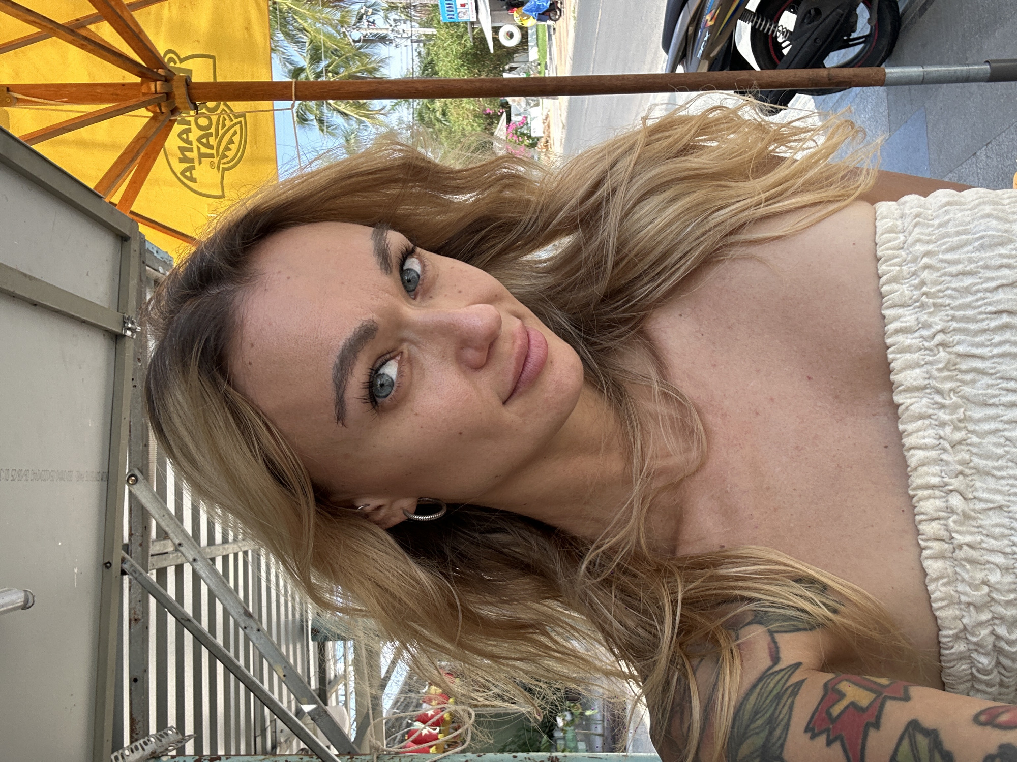 A woman with blonde hair and light eyes is looking at the camera. She is wearing a white, textured top and has a tattoo on her left arm. She appears to be seated under a yellow umbrella, next to a metal structure. The background reveals a street scene with a motorbike, palm trees, and buildings, indicating an urban environment. Based on the provided information, the location is Phu Thuy Ward, Vietnam.