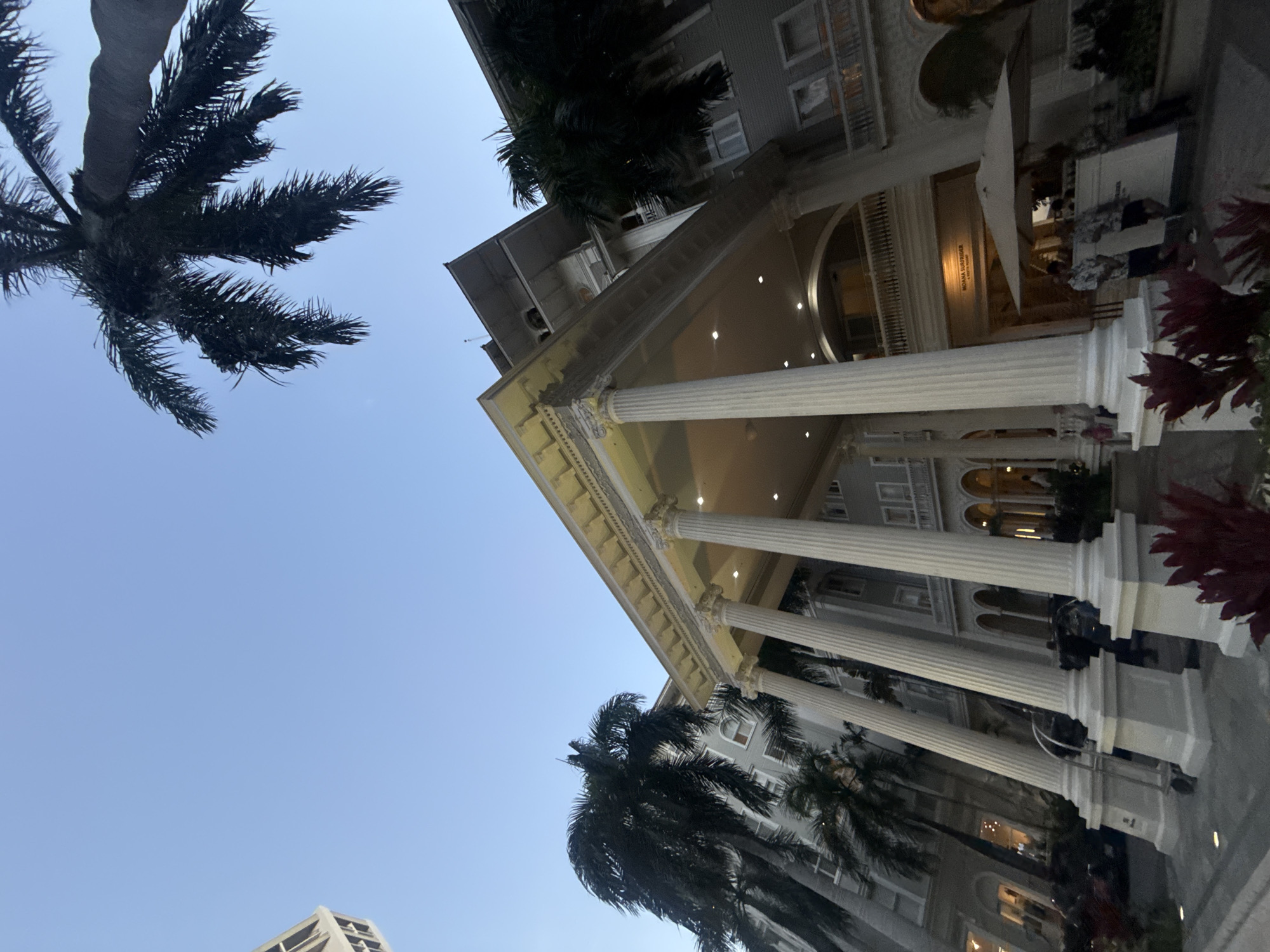 The image depicts the Moana Surfrider, A Westin Resort & Spa, Waikiki Beach, in Honolulu, United States of America, viewed from a low angle. Palm trees frame the sky and the building, with classical-style columns supporting an elaborate pediment. Some people are visible inside the building.