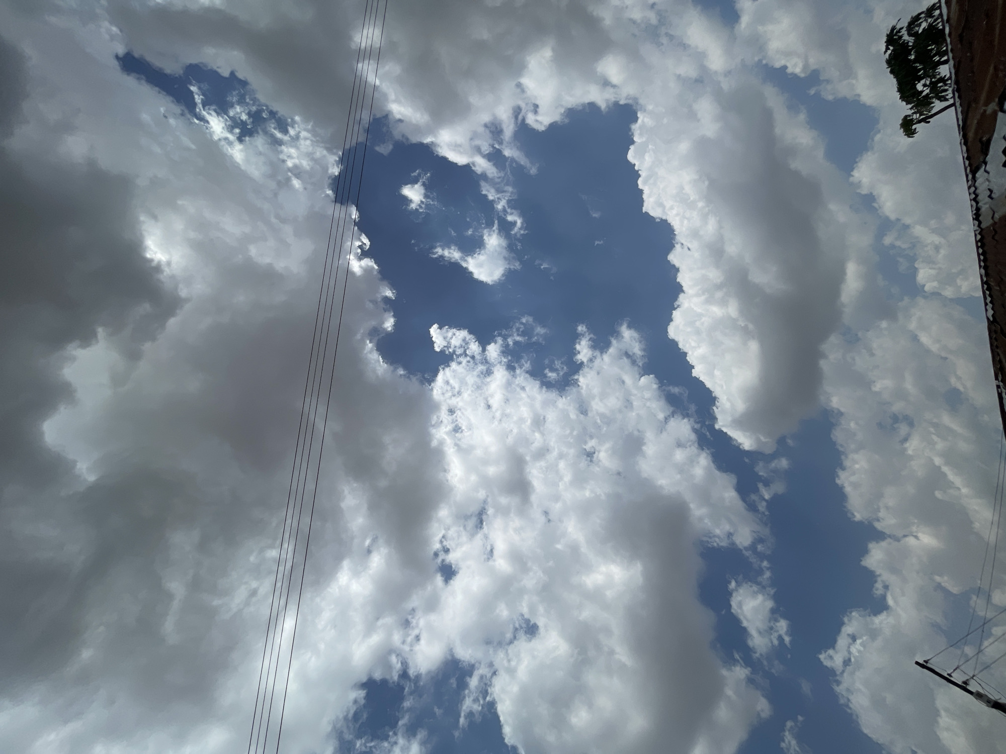 The image is an upward-facing shot of a sky filled with cumulus clouds, with patches of blue sky visible between them. Some clouds are bright white, while others are gray, suggesting varying densities and depths. Electrical power lines are visible on the left side of the image. A tree and building are present on the right side of the image. The location is Jos, Nigeria.