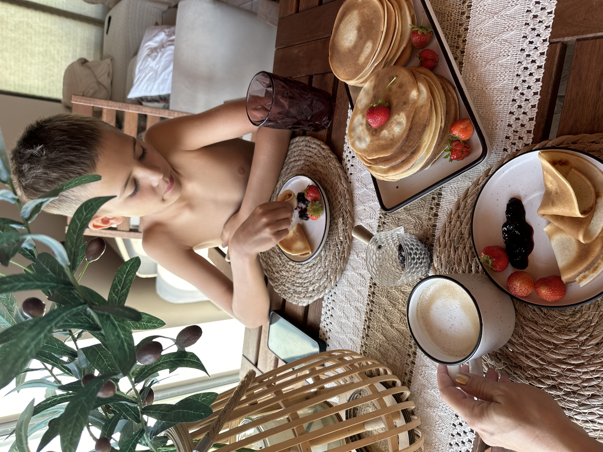 A young boy with dark, short hair, shirtless, sits at a wooden table and eats pancakes. He is looking down at his plate with his eyes closed, and his expression appears content. He is holding a fork in his right hand, and has a small plate of pancakes, strawberries, and jam in front of him. To his right is a tall glass of dark liquid, a stack of pancakes on a tray, and a coffee cup with latte art. The scene is set outdoors on a sunny day, as indicated by the bright lighting and the presence of what appears to be an olive tree branch in the foreground. The background shows a blurred interior with furniture. The overall mood is relaxed and leisurely, suggesting a breakfast or brunch setting.