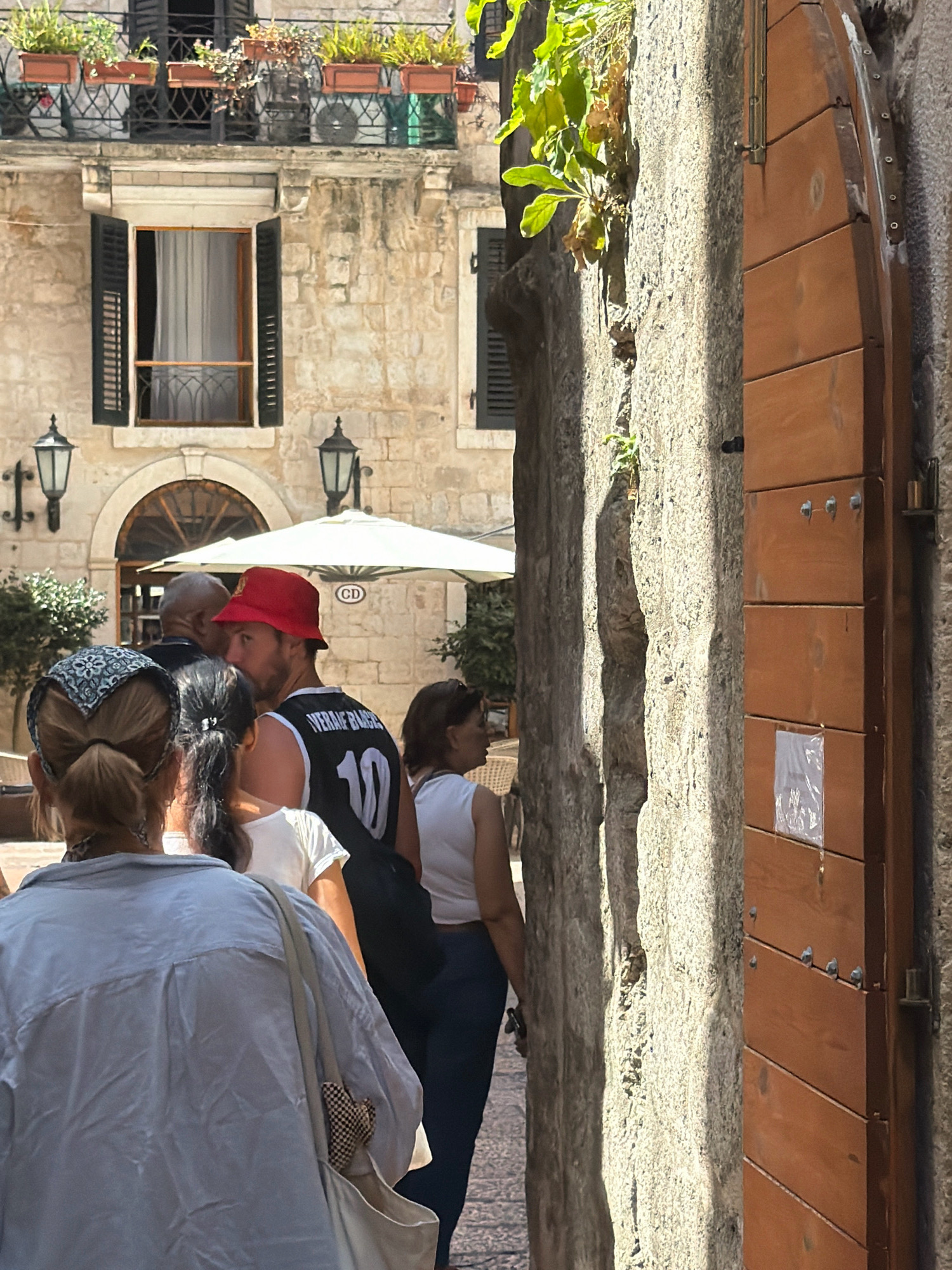 In Kotor, Montenegro, a group of people are walking down a narrow street. In the foreground, a person wearing a light blue shirt walks away from the camera, with a tote bag slung over their shoulder. To their right, another person is seen from the back, wearing a black tank top with the number "10" and the name "VERME BADGE" visible on it. They are also wearing a red bucket hat. Behind them, a woman in a white top walks away. The street is paved with cobblestones. The buildings on either side are made of stone, with traditional shutters and balconies. A large wooden door is visible on the right side of the image. The scene is brightly lit by the sun, casting shadows.
