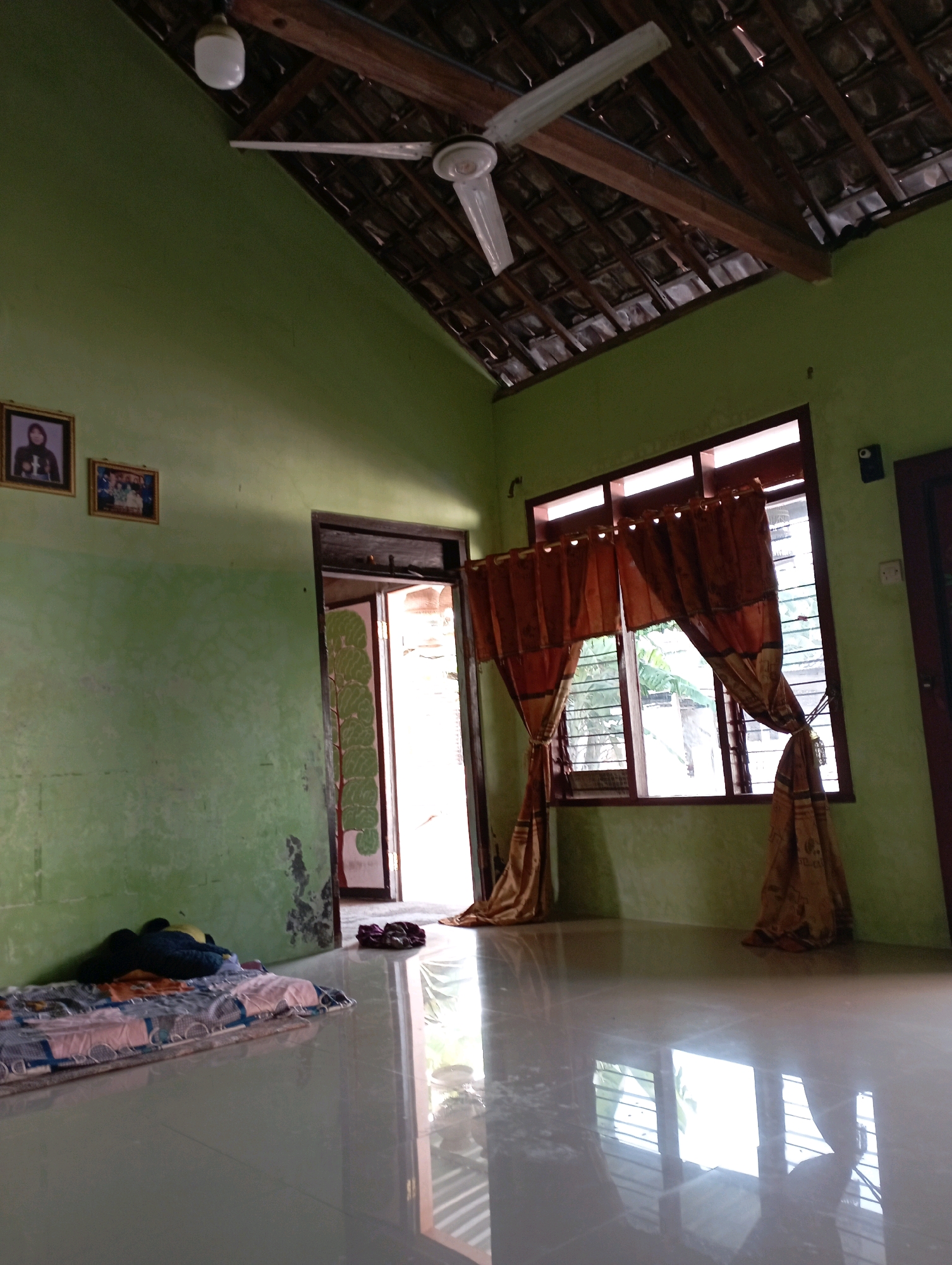 The image depicts a room with light green walls and a tiled floor that reflects the surroundings. In the upper portion of the image, exposed wooden roof beams are visible, supporting a tiled roof. A white ceiling fan with three blades is mounted centrally on the ceiling. To the left, on the wall, hang two framed pictures. One frame contains a portrait of a person wearing a headscarf, and the other contains a group photograph. Near the floor on the left side of the room, a mattress with patterned bedding lies on the floor, with dark-colored stuffed toys placed on it. To the right of the mattress, a doorway leads outside, revealing an open door with a mural of a tree painted on the inner side of the door. Outside the doorway, a pair of sandals rests on the floor. Further to the right, a large window is covered by a set of light brown, patterned curtains. The window overlooks an exterior with horizontal blinds and foliage. The floor is highly reflective, showing distorted reflections of the window and the ceiling fan. The overall impression is of a simple, lived-in domestic space. The provided contextual information specifies the location as Sonobekel, Indonesia.