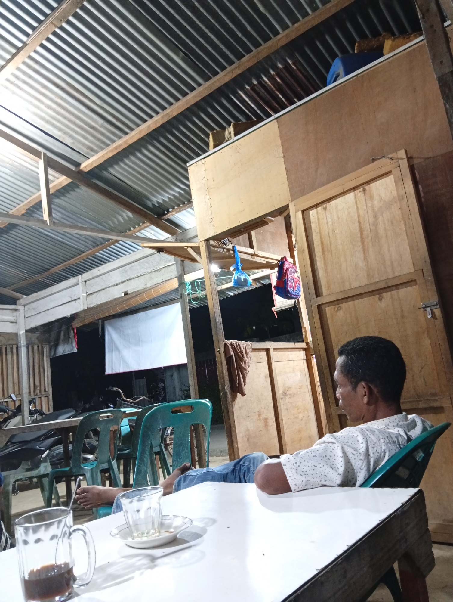 The image depicts an indoor scene at what appears to be a simple, possibly open-air establishment in Lhokseumawe, Indonesia. A man, wearing a patterned shirt and jeans, is seated in a green plastic chair, looking away from the camera. He is at a white table, on which two glasses and a saucer are visible. One glass contains a dark liquid, likely coffee. In the background, there are other green plastic chairs and tables, and a motorcycle is partially visible. The ceiling is made of corrugated metal sheets supported by wooden beams. The walls are constructed from wooden planks and plywood. A white sheet hangs on the wall in the mid-ground. A blue plastic container and some fabric items are visible on a shelf above a wooden structure. The overall impression is of a casual, everyday setting, possibly a local eatery or rest stop.