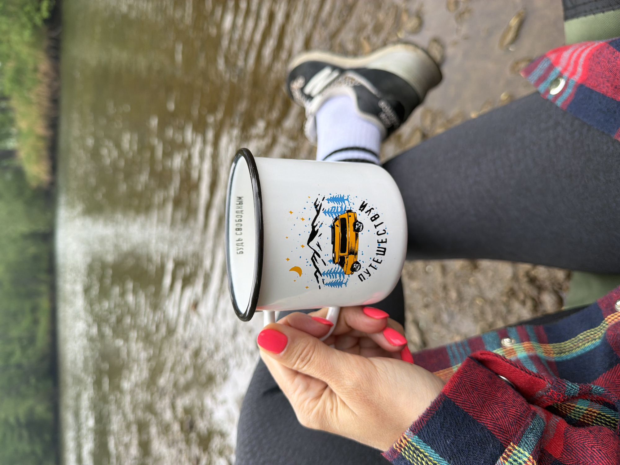 A person wearing black leggings, a red and blue plaid shirt, white socks, and black and white sneakers is sitting near a body of water. They are holding a white enamel mug with a black rim and an illustration of a yellow vehicle, blue trees, and stars. The mug also has writing on it in a language other than English. The nails of the person holding the mug are painted pink. The water appears to be a stream or river with a muddy bottom and vegetation on the bank in the background. The location is not identified as Trek, Russia in the image.