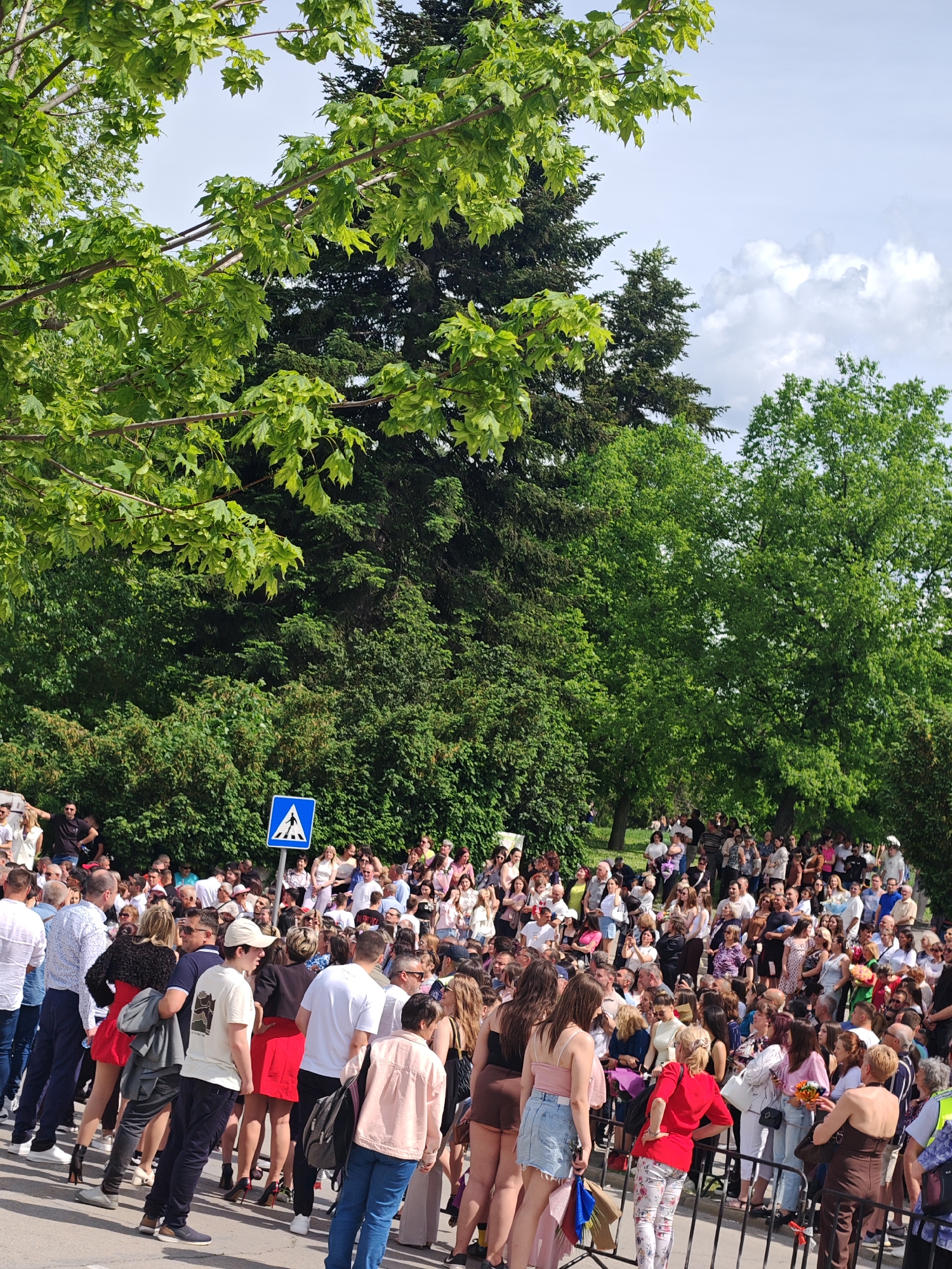 The image shows a large crowd of people gathered outdoors in what appears to be a park-like setting in Shumen, Bulgaria. Many trees are in the background, and the people are closely packed together, appearing to be waiting or observing something. There is a pedestrian crossing sign. Some people are dressed in casual attire. A metal barricade is set up in the foreground, separating the crowd from an area.