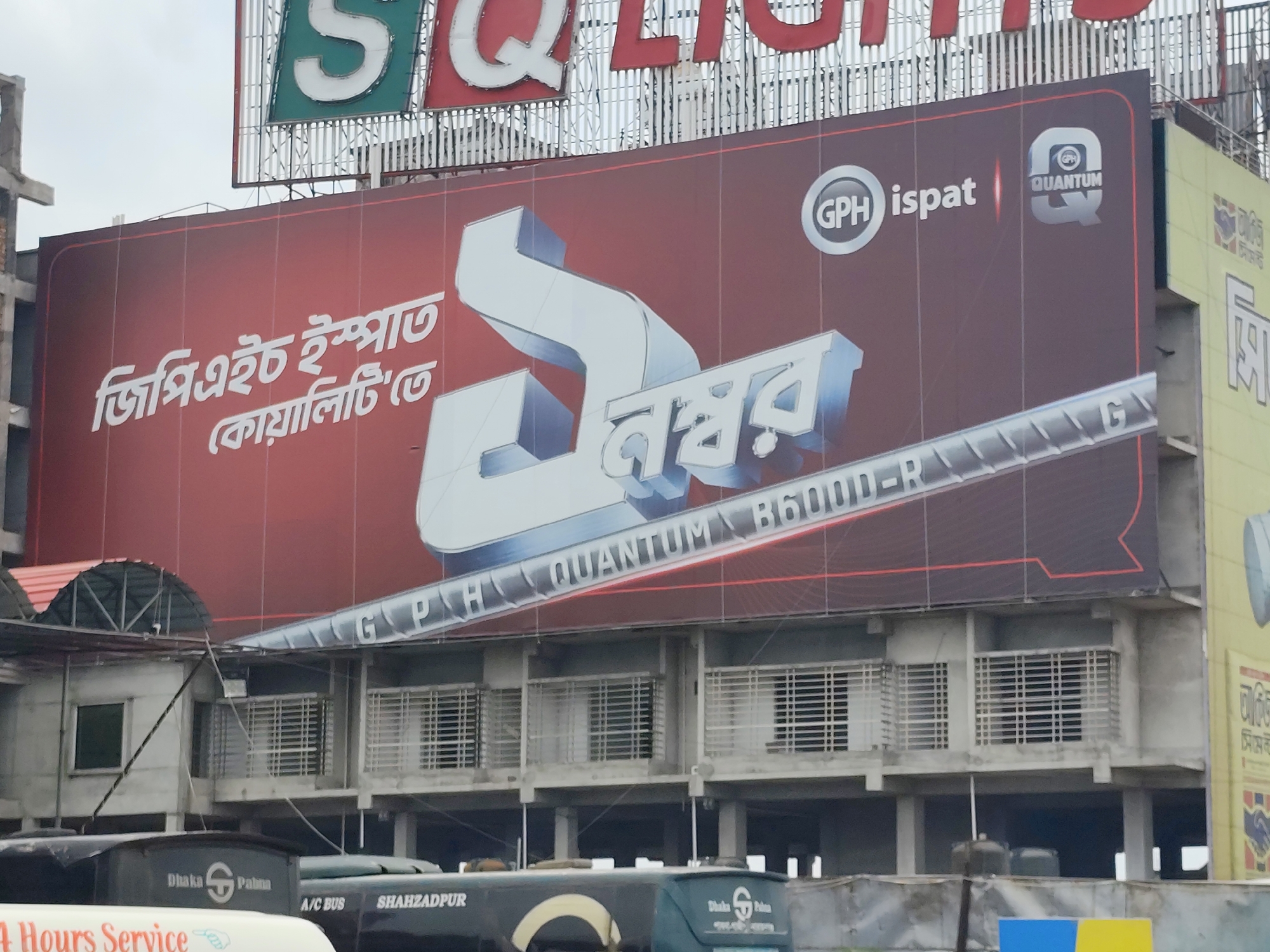The image captures an urban scene, likely in Hatikumrul, Bangladesh, featuring a large billboard advertisement dominating the view. The advertisement is for "GPH Ispat" and "GPH Quantum B6000-R," showcasing a stylized representation of the number "1" in Bengali script.
Below the billboard is a building with multiple levels and barred windows. Several buses are parked in front of the building. Some buses have the word "Dhaka," followed by an image, and the word "Palma." Another bus has the words "A/C BUS SHAHZADPUR." The background includes overcast skies.