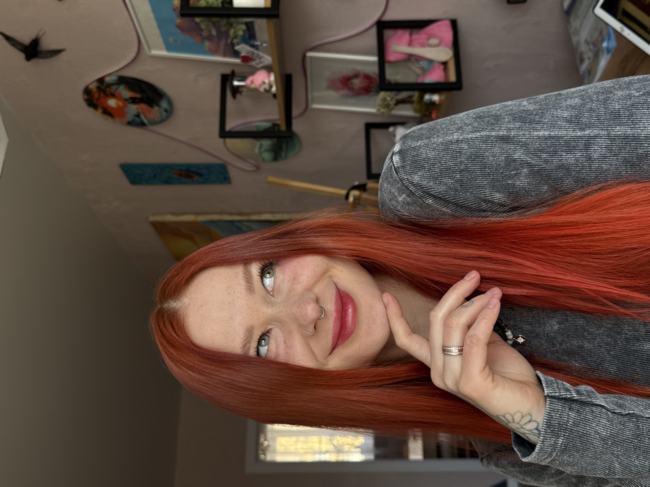 The image displays an indoor, close-up portrait of a female individual situated in Rostov-on-Don, Russia. The subject, positioned centrally, possesses long, straight, vibrant red hair with a subtle off-center part. She has fair skin exhibiting freckles across her nose and cheeks, and her light green eyes are directed upwards and to the viewer's right. Her lips are closed in a slight smile, accentuated by pinkish-red lip gloss, and a silver septum ring is visible in her nose. Her right hand is raised, with her chin lightly resting on her index finger while her thumb supports her jawline. A silver band-style ring with a textured pattern is present on her right ring finger, and a black-ink daisy tattoo is visible on the inner side of her right wrist. She is attired in a grey, long-sleeved shirt with a ribbed texture, and a dark beaded necklace with a light-colored pendant is visible around her neck.
The background features a light pink wall, illuminated by natural light entering from a window or doorway visible on the far left. The wall is decorated with multiple items: a black silhouette of a bird, two rectangular black floating shelves holding various objects including a pink hourglass, a small pink pig figurine, a framed pink artwork, a small plant, and a framed pink teddy bear holding an elongated light-colored object. Circular and rectangular wall art pieces, including a sunset scene and a small blue artwork, are also affixed to the wall. Decorative thin, wavy pink lines, possibly electrical wires, are routed across the wall between the shelves and art. A large painting with warm tones is partially visible on the upper left side of the decorated wall section.