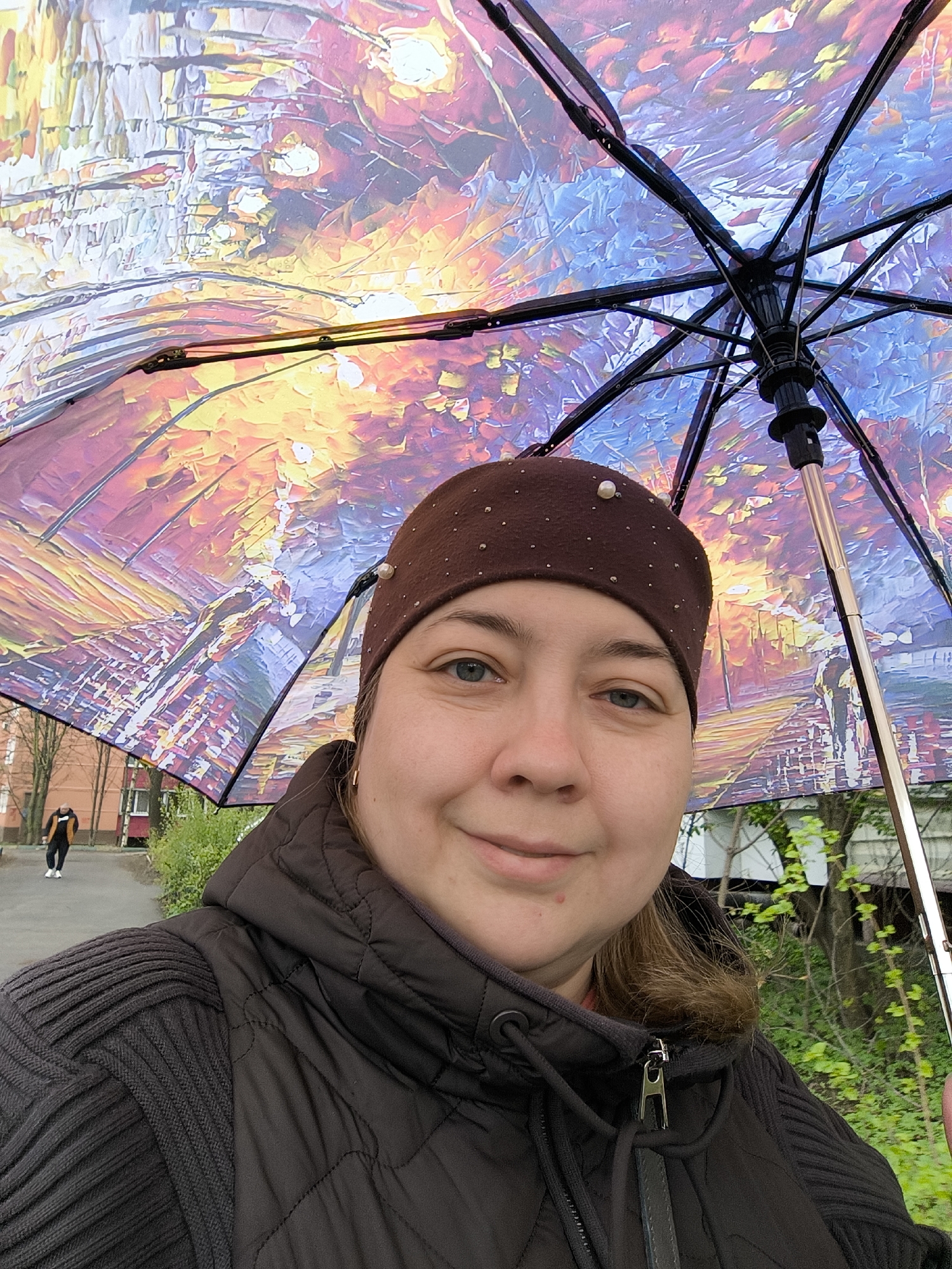 A woman is taking a selfie while holding an umbrella. She is wearing a brown beanie with small white pearls, a dark quilted jacket with a sweater underneath. The umbrella has an artistic, colorful design. In the background, there is a paved pathway, a building, trees, and another person walking in the distance. Based on visual cues and metadata, this photo was taken in Khimki, Russia.