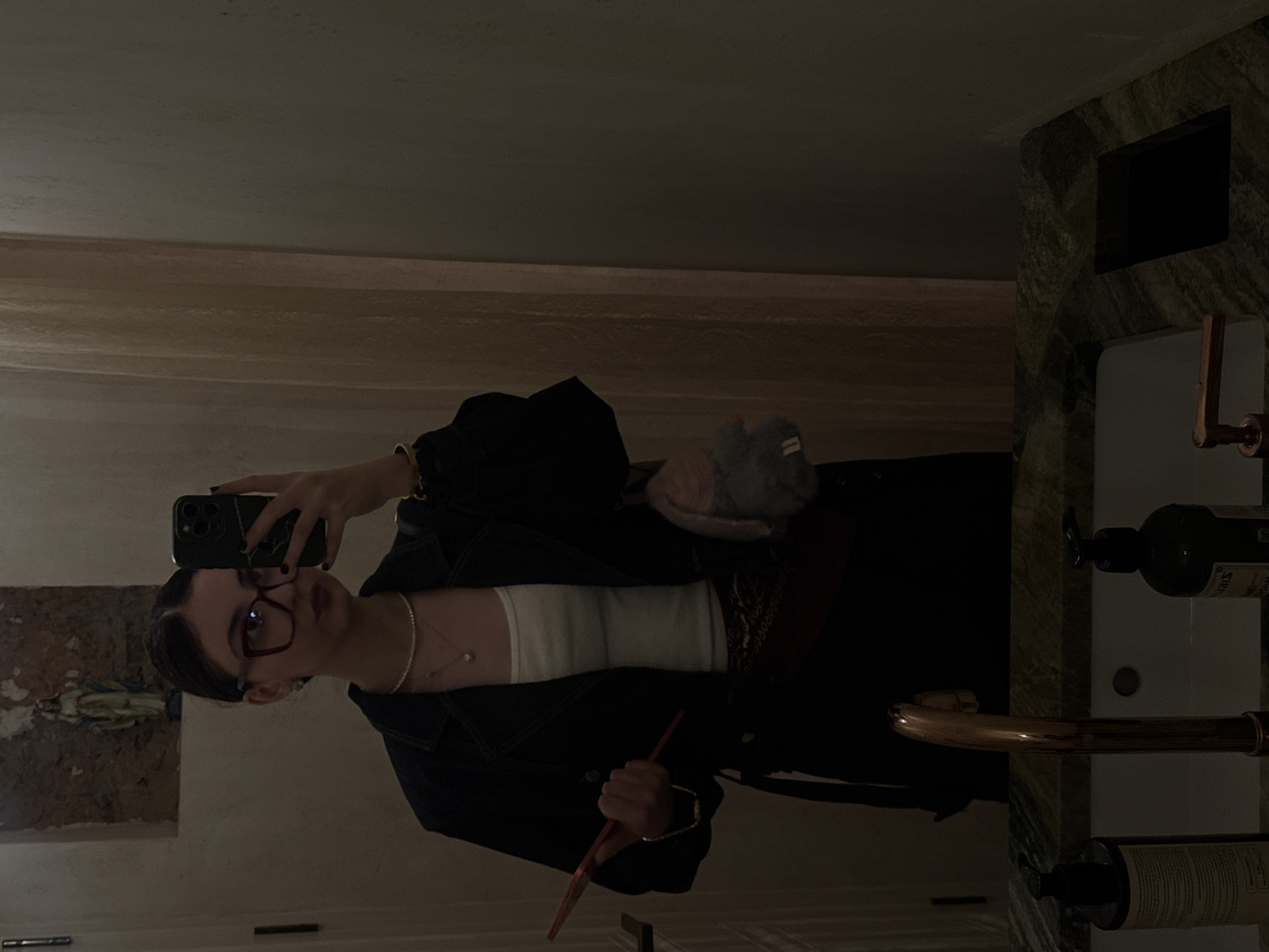 A woman is taking a selfie in a bathroom. She is wearing glasses, a white top, a dark jacket, and jewelry (necklace and bracelets). She is holding a phone in one hand and a red object in the other. There is a sink and countertop visible, with various bottles and items arranged on it. The bathroom has a marble or stone aesthetic. There is a decorative element on the wall in the background. There is no information in the photo that allows the location to be confirmed as Yerevan, Armenia.