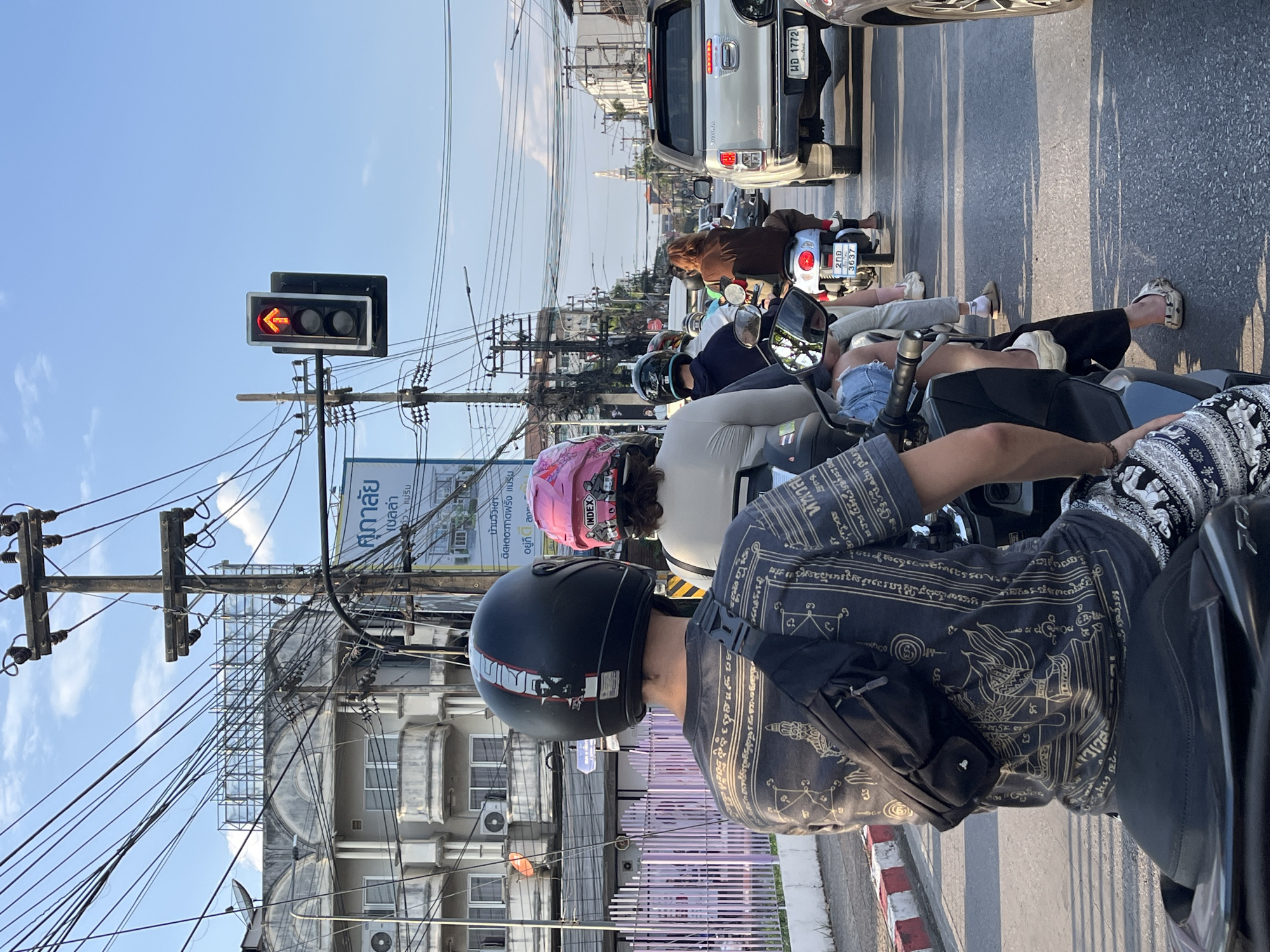 The image shows a street scene in Chiang Mai City Municipality, Thailand, at a traffic light intersection. Multiple motorbikes are stopped, waiting for the light to change. The perspective is from a rider on one of the motorbikes. A red left-turn arrow traffic signal is visible. In front of the motorbikes is a white pickup truck. Various buildings, telephone poles, and a billboard are in the background, cluttered with electrical wires. The billboard has Thai script and the words "ศุภาลัย" and "INDEX" are legible. The weather appears to be sunny.