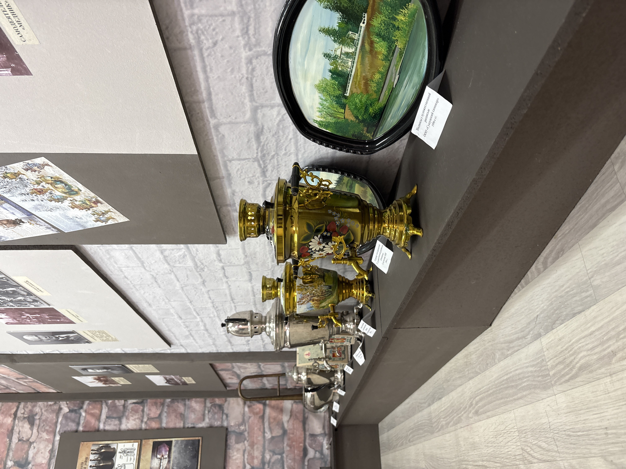 The image shows a museum display in Suksun, Russia, featuring several antique samovars and decorative trays. The items are arranged on a dark shelf against a white brick wall. In the background, there are framed photographs and artwork. The floor is light-colored laminate. The lighting is even, suggesting an indoor museum setting.