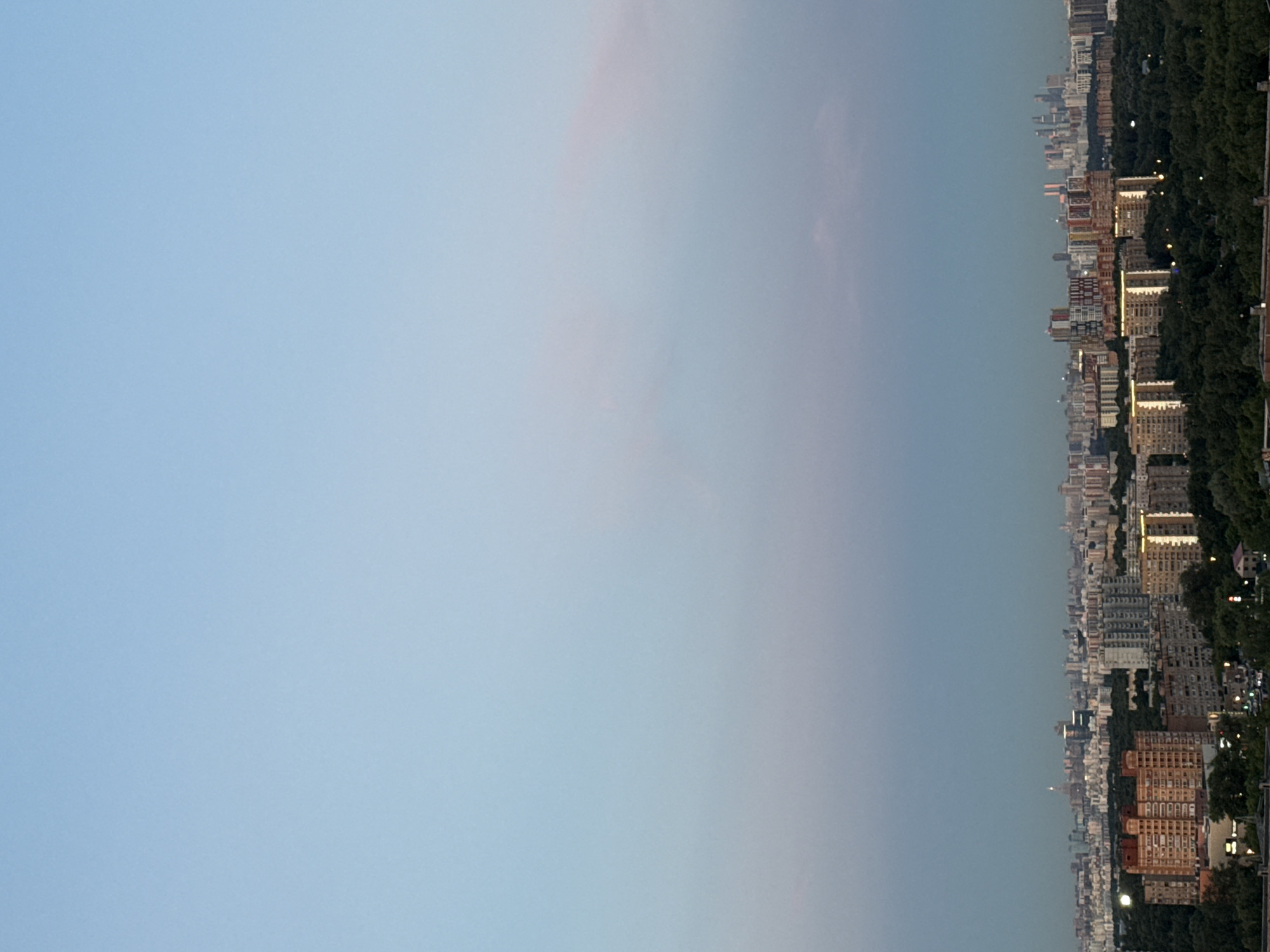 This is a vertical, full-frame, long-distance photograph of a cityscape at dusk. The image is split roughly in half horizontally. The left side of the image shows a pale blue sky with subtle pinkish hues, suggesting the fading light of sunset or the transition to twilight. The right side of the image depicts a dense urban landscape with numerous buildings of varying heights and architectural styles.
The city appears to be situated along a tree-lined area, possibly a park or forest, which forms a dark green band along the edge of the urban sprawl. Many of the buildings are illuminated with warm yellow lights, indicating that the activity within them continues as evening sets in. The buildings are packed closely together, showcasing a dense urban environment. Some of the buildings are lighter in color, while others are brick-red, adding variety to the scene. The overall impression is of a large, active city as day transitions into night. The location is identified as Khimki, Russia, and the time of day is dusk. There is no visible text in the image.